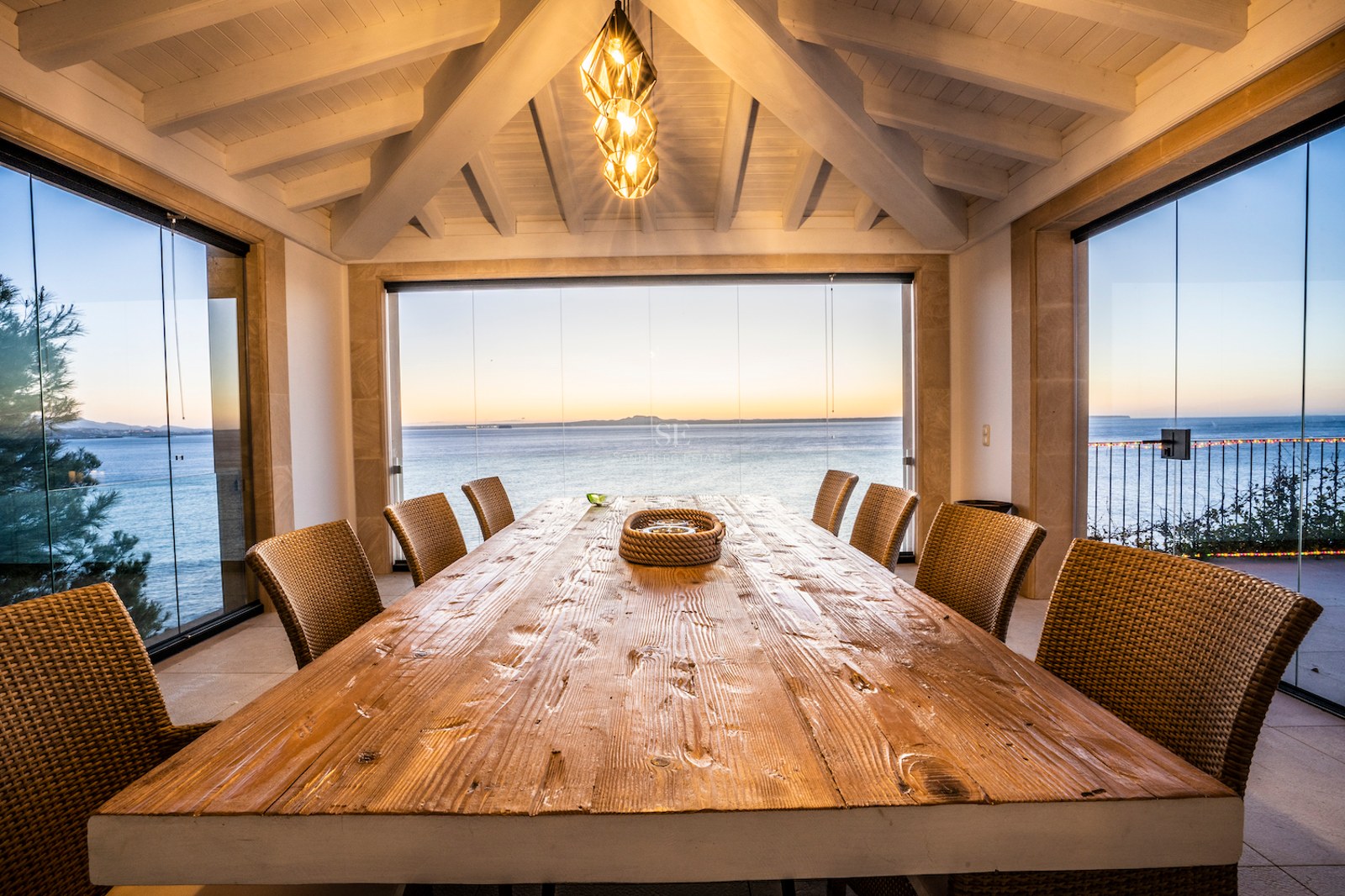Large wooden dining table on a glass-enclosed terrace with sunset views over the Mediterranean sea.