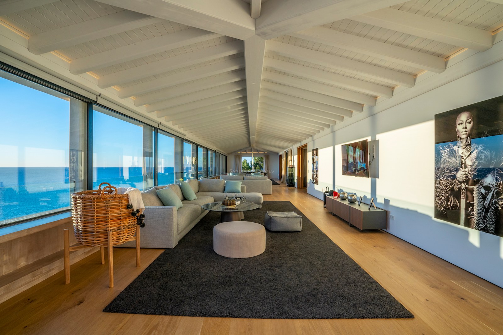 Contemporary living room with white beamed ceilings, oak floors, and floor-to-ceiling windows overlooking the sea.