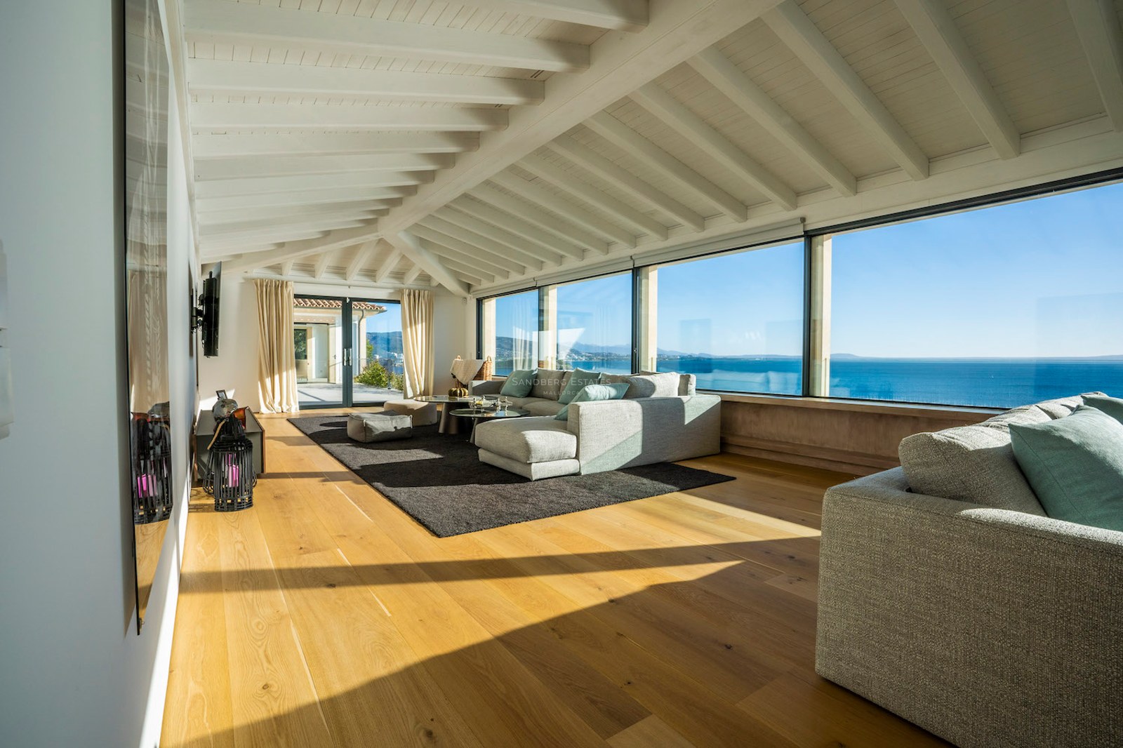 Bright living room featuring large panoramic windows overlooking the sea, white beamed ceilings, and oak wood floors.