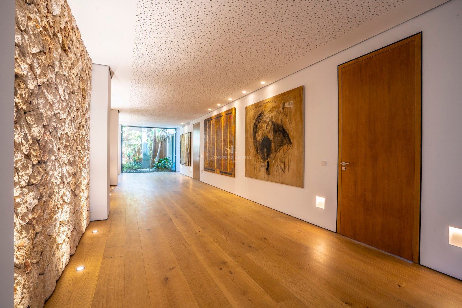A long modern hallway with wide oak floorboards, white walls with art, and a glass door leading to a garden.