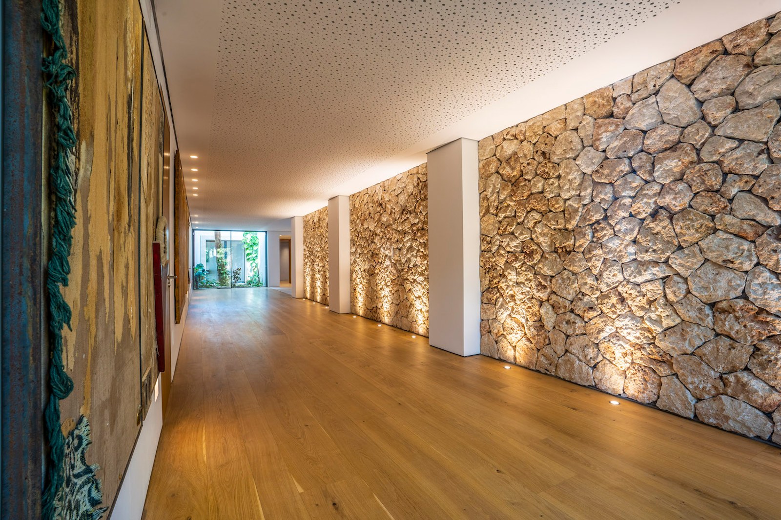 Modern interior hallway featuring a textured stone wall, recessed floor lighting, and light oak wood flooring.