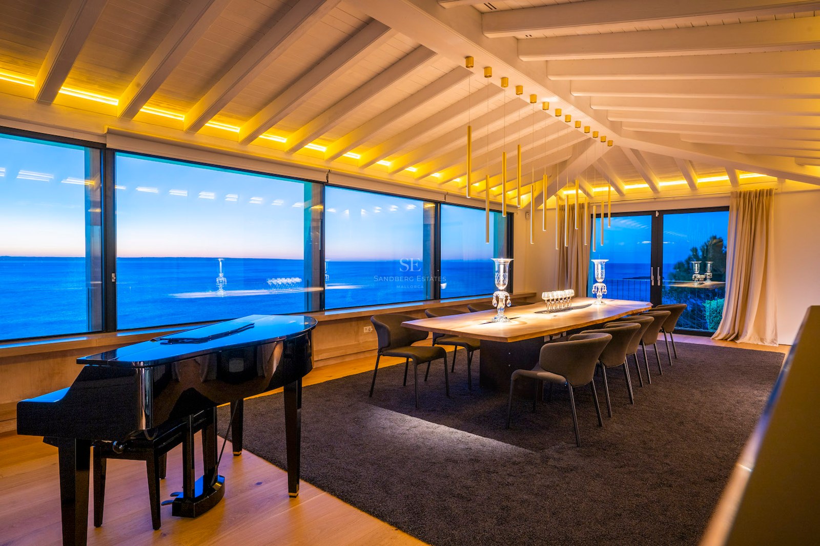A modern dining room featuring a grand piano, large wooden table, and floor-to-ceiling windows overlooking the sea at dusk.