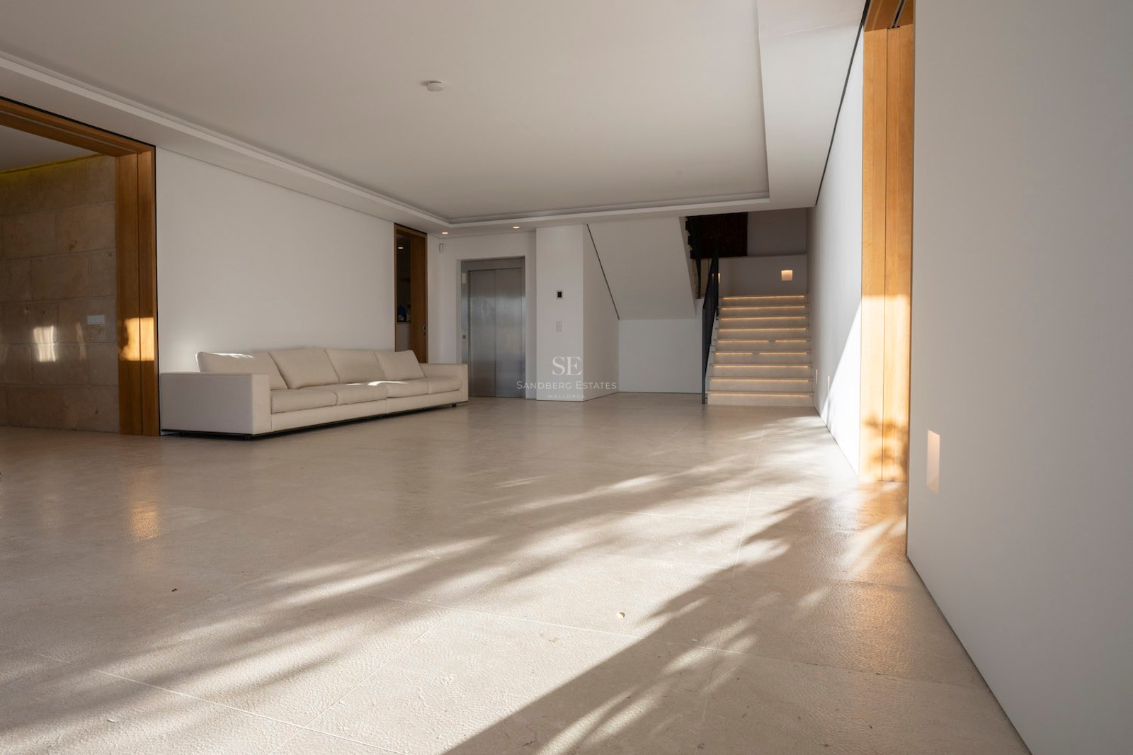 Spacious minimalist hall with cream stone floors, a white sofa, a private elevator, and a stone staircase.