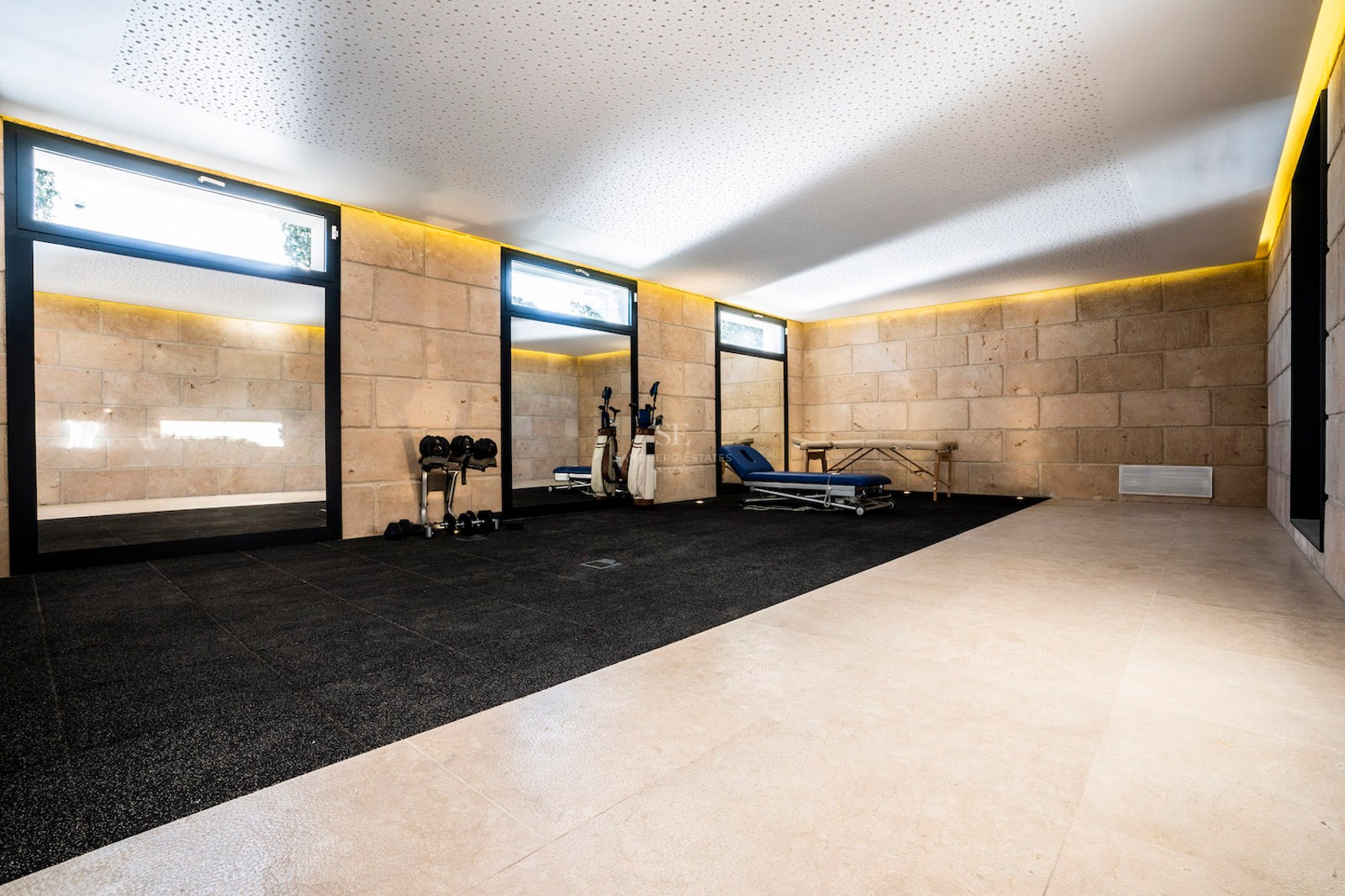 Modern home gym featuring natural stone walls, professional rubber flooring, a massage table, and indirect ceiling lighting.