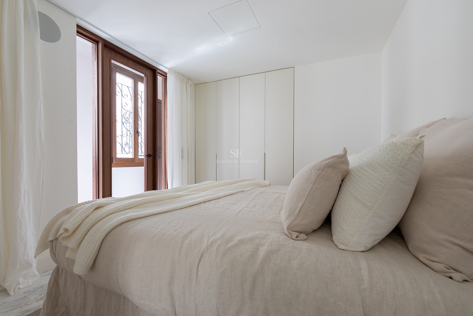 Dormitorio luminoso con ropa de cama de lino beige, armarios blancos minimalistas y puerta de madera al balcón.