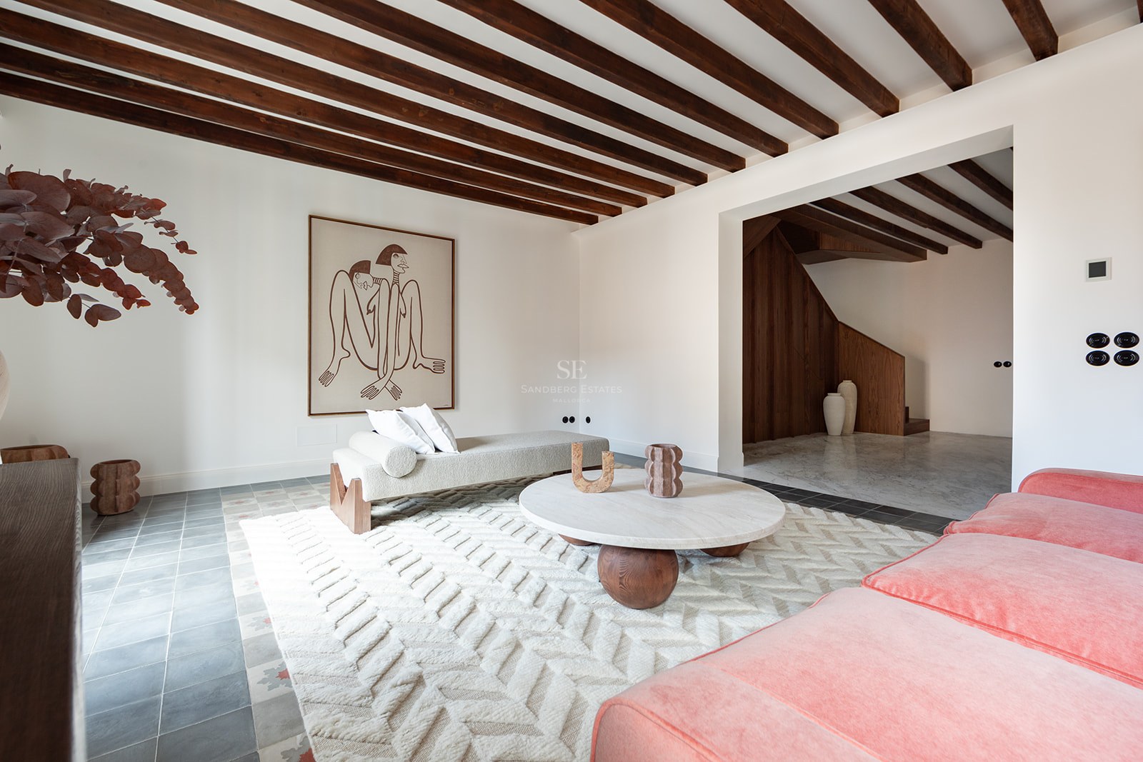 Salón moderno con vigas de madera oscura, sofá de terciopelo terracota, mesa de mármol y suelo de baldosa artesanal.