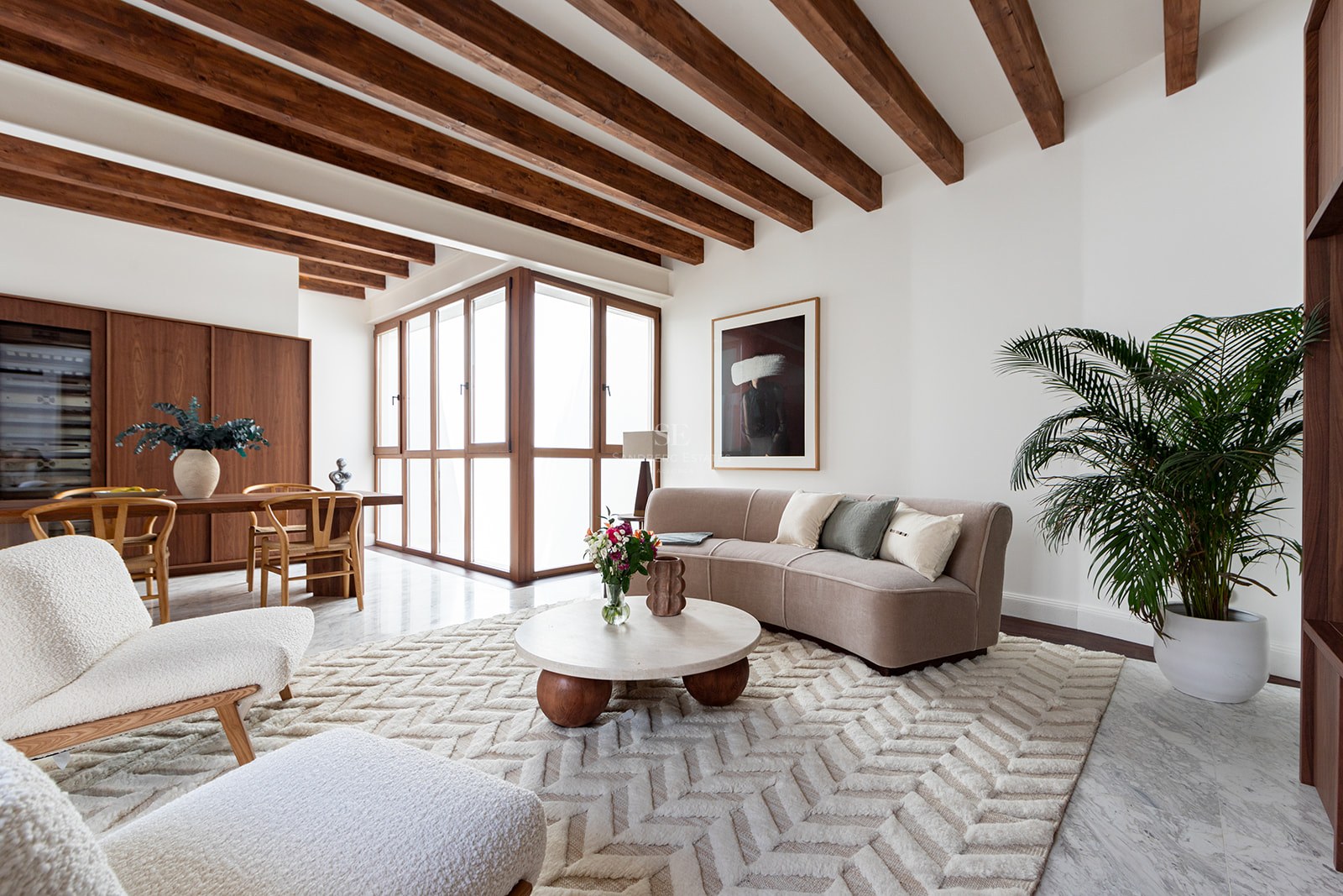 Sala de estar moderna con vigas de madera oscura, suelo de mármol, sofá beige curvo y sillones de borreguillo blanco.