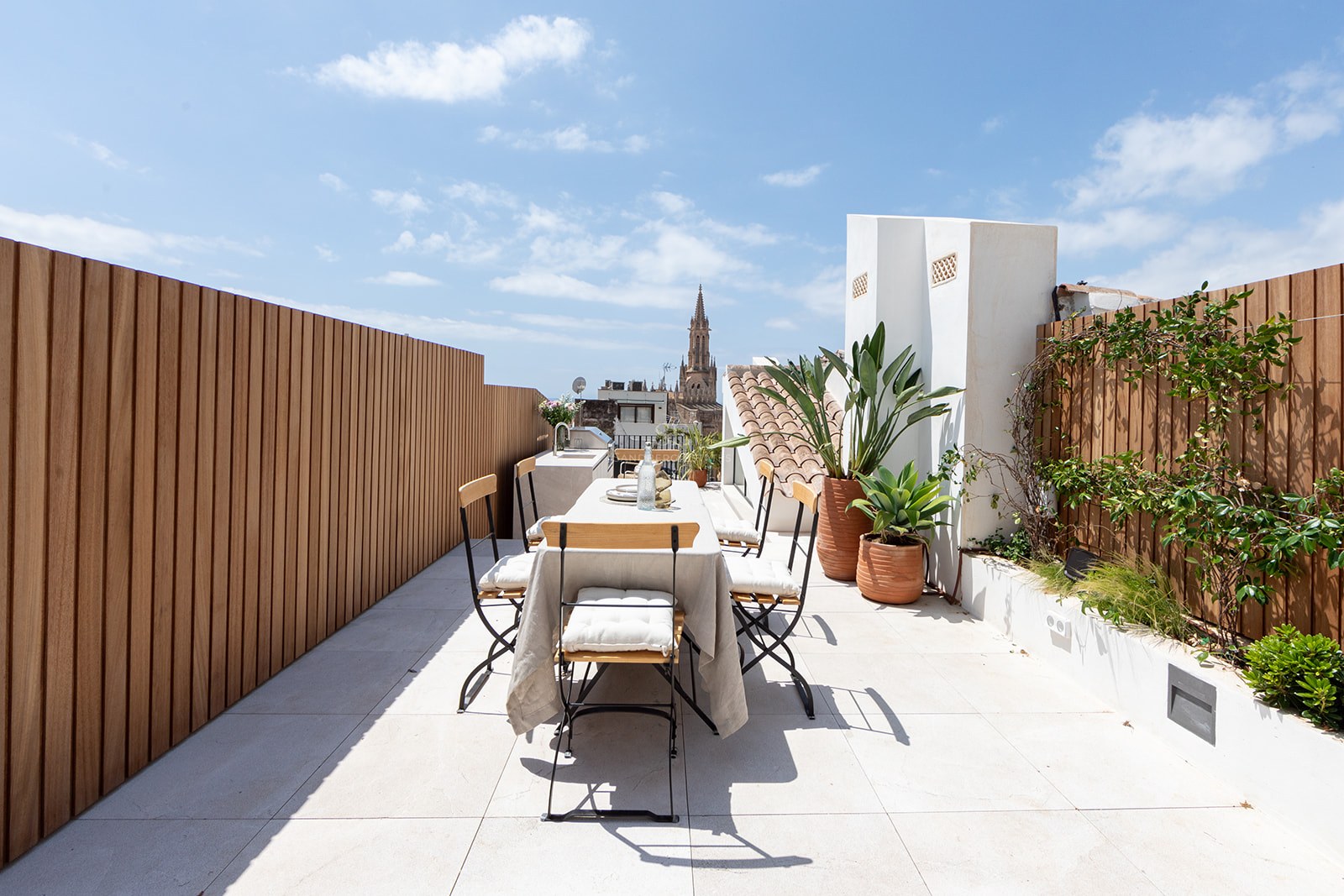 Terraza moderna con mesa de comedor, cercado de madera y vistas a la Catedral de Palma bajo un cielo azul despejado.