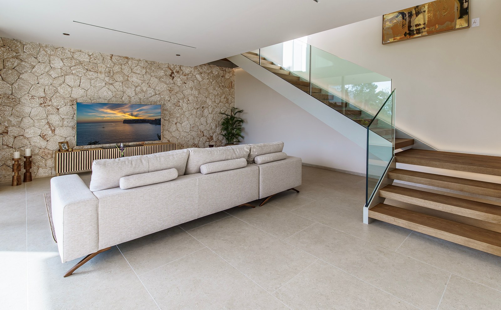 Salon spacieux avec canapé beige, mur en pierre décoratif, télévision et escalier en bois avec garde-corps en verre.