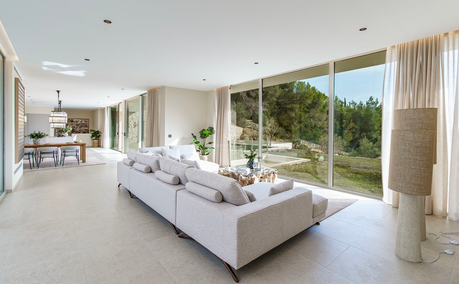 Grand salon moderne avec un canapé gris et des baies vitrées donnant sur une forêt verdoyante.