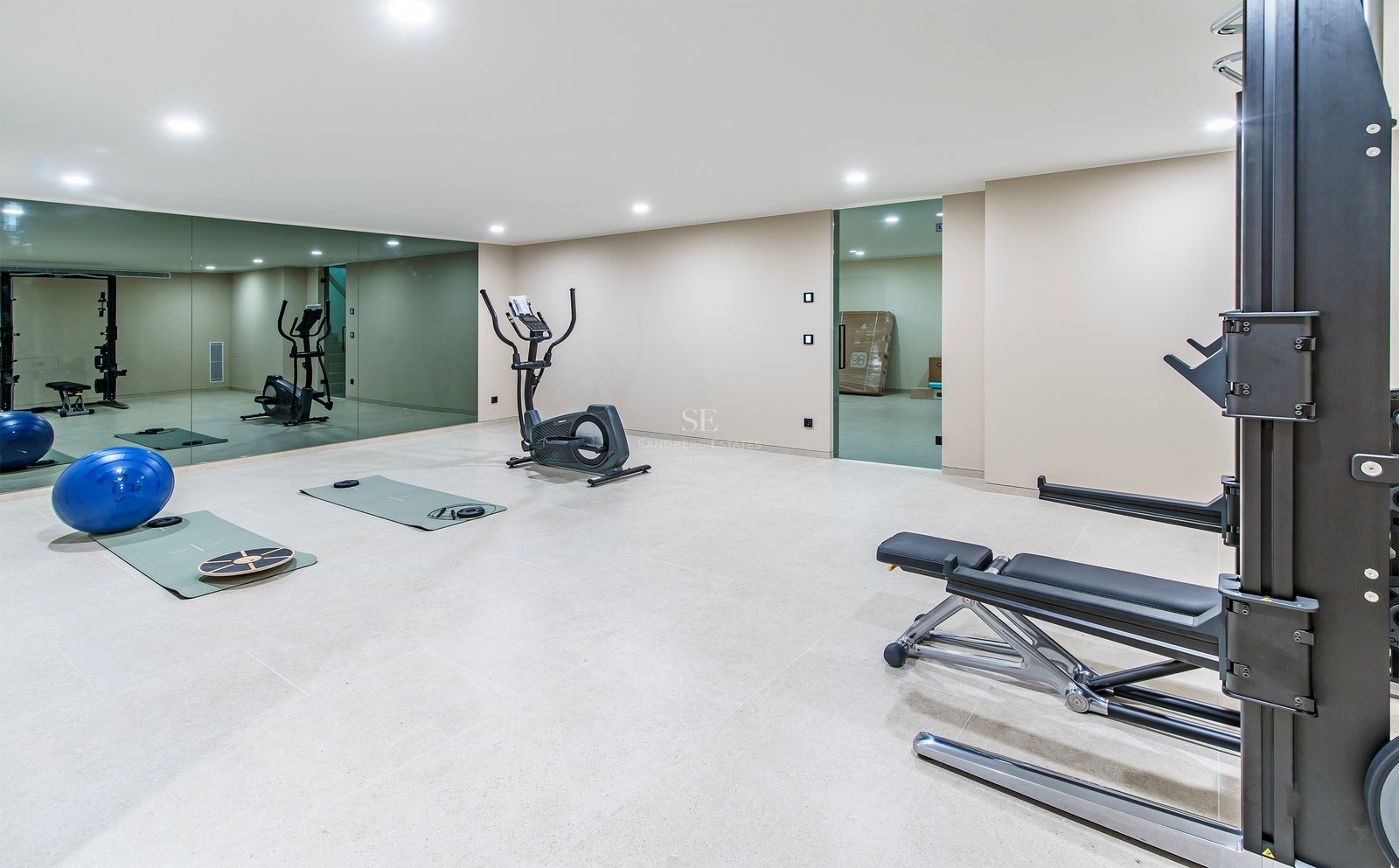 Salle de sport avec vélo elliptique, banc de musculation et grands miroirs sous des plafonniers brillants.