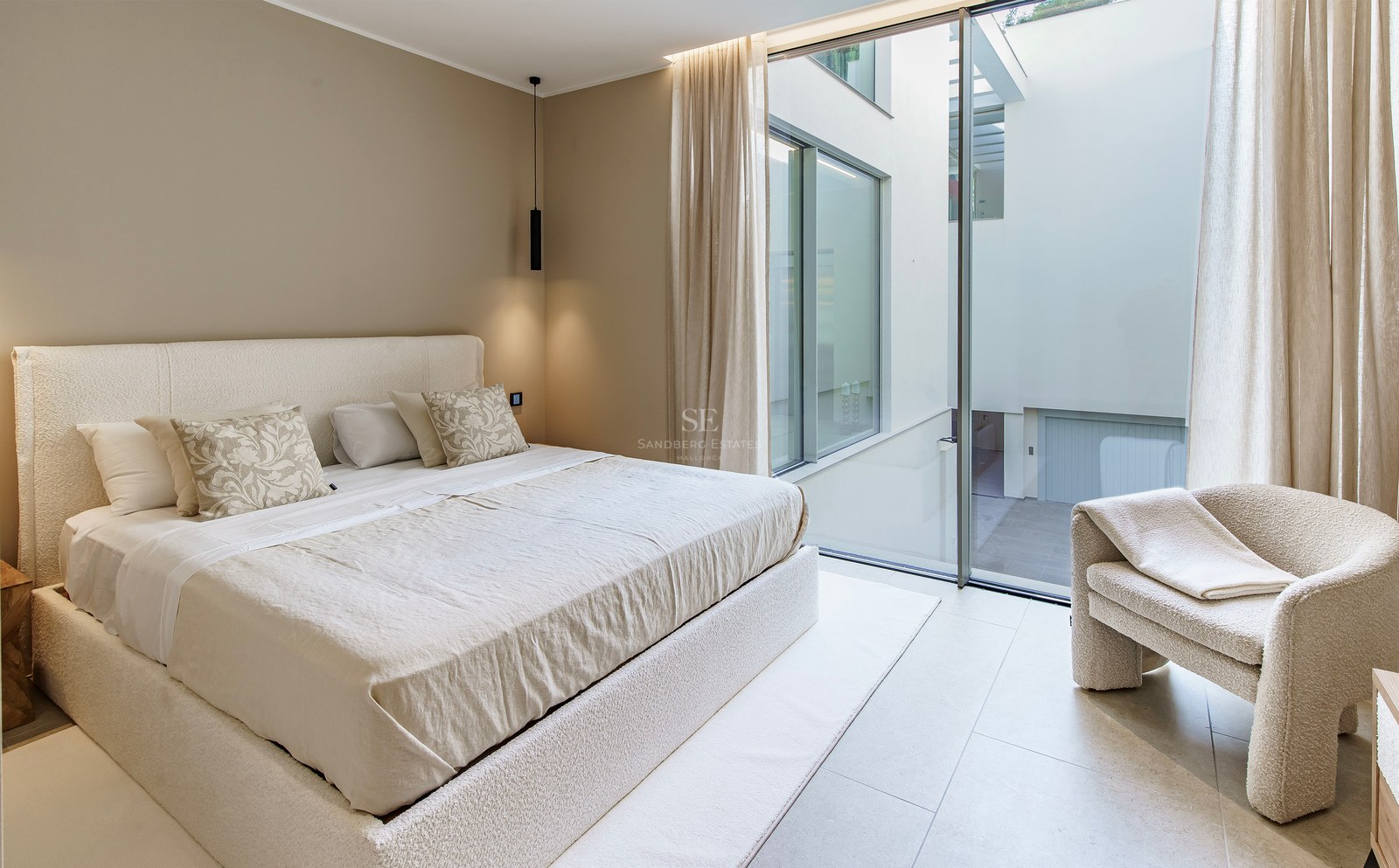 Chambre minimaliste avec lit en bouclé crème, fauteuil assorti et grandes baies vitrées donnant sur un patio intérieur.