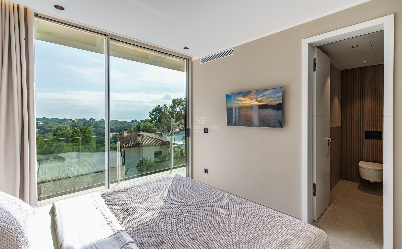 Une chambre moderne dotée d'une grande baie vitrée donnant sur une forêt et la mer, avec une salle de bain attenante.