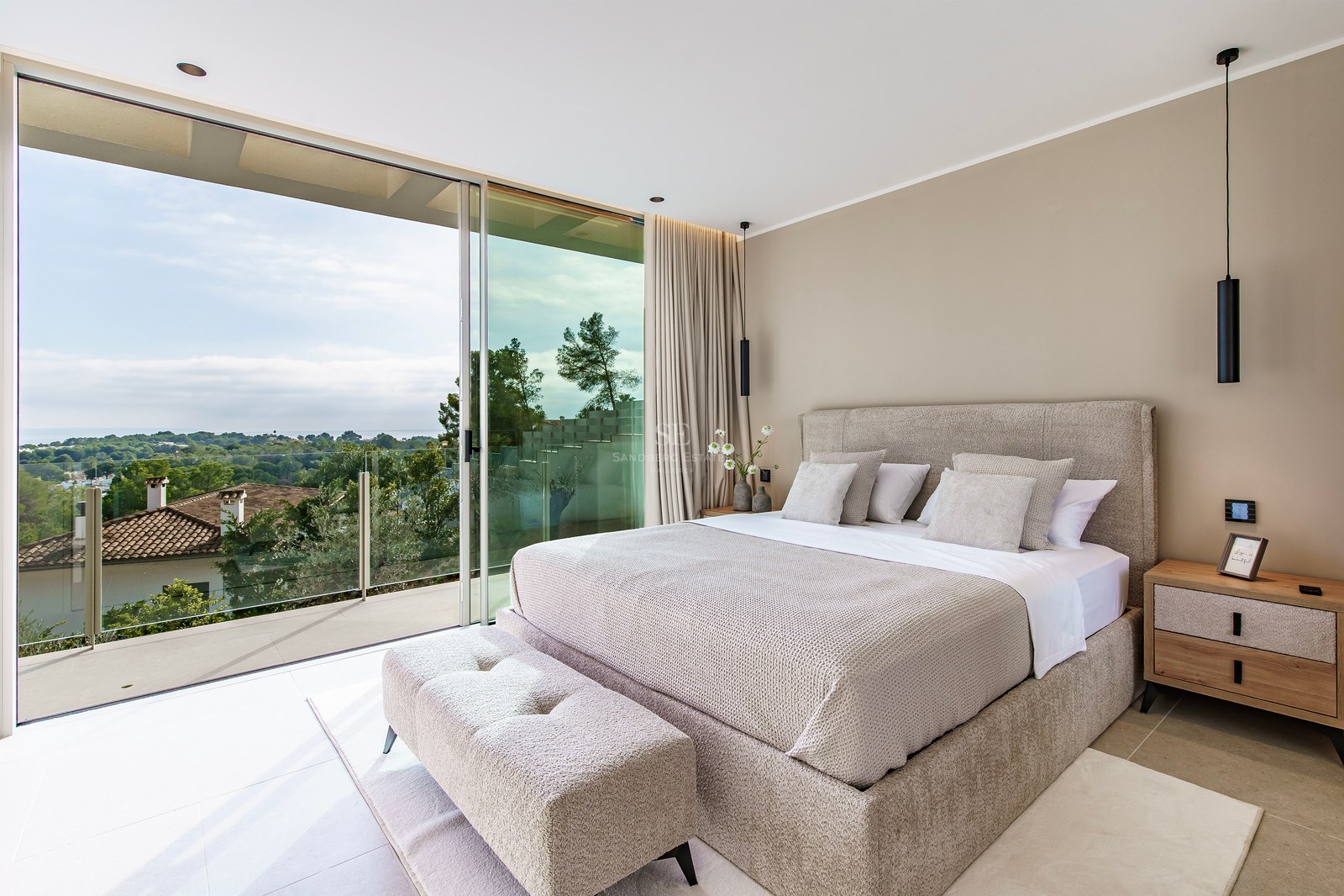 Chambre de luxe avec lit beige et grandes baies vitrées ouvrant sur un balcon avec vue sur la verdure.