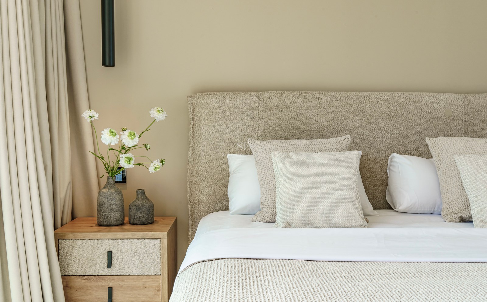 Lit de luxe avec tête de lit texturée, table de chevet en bois et vases décoratifs avec fleurs blanches.
