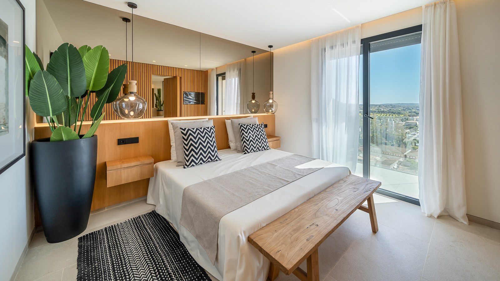 Chambre lumineuse avec tête de lit en bois, grand miroir, suspensions en verre et accès au balcon.