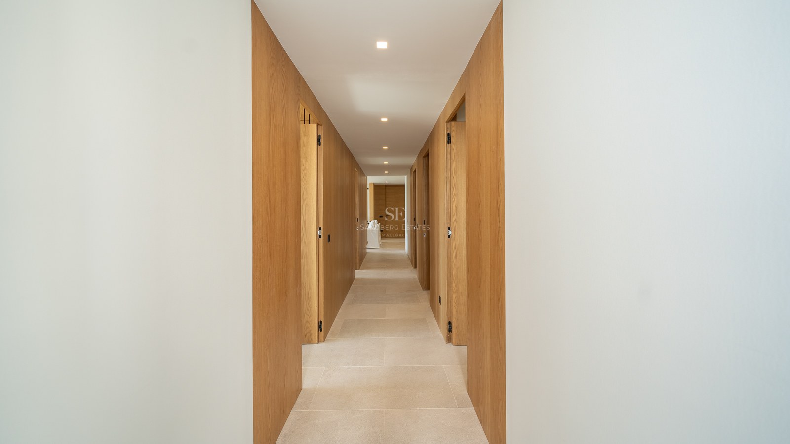 Long couloir moderne avec murs en bois clair, portes invisibles intégrées et sol en pierre beige sous spots LED.