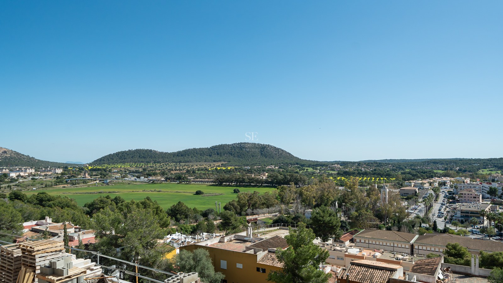 Vue en plongée d'une vallée verdoyante, de montagnes lointaines et d'un village méditerranéen sous un ciel bleu clair.