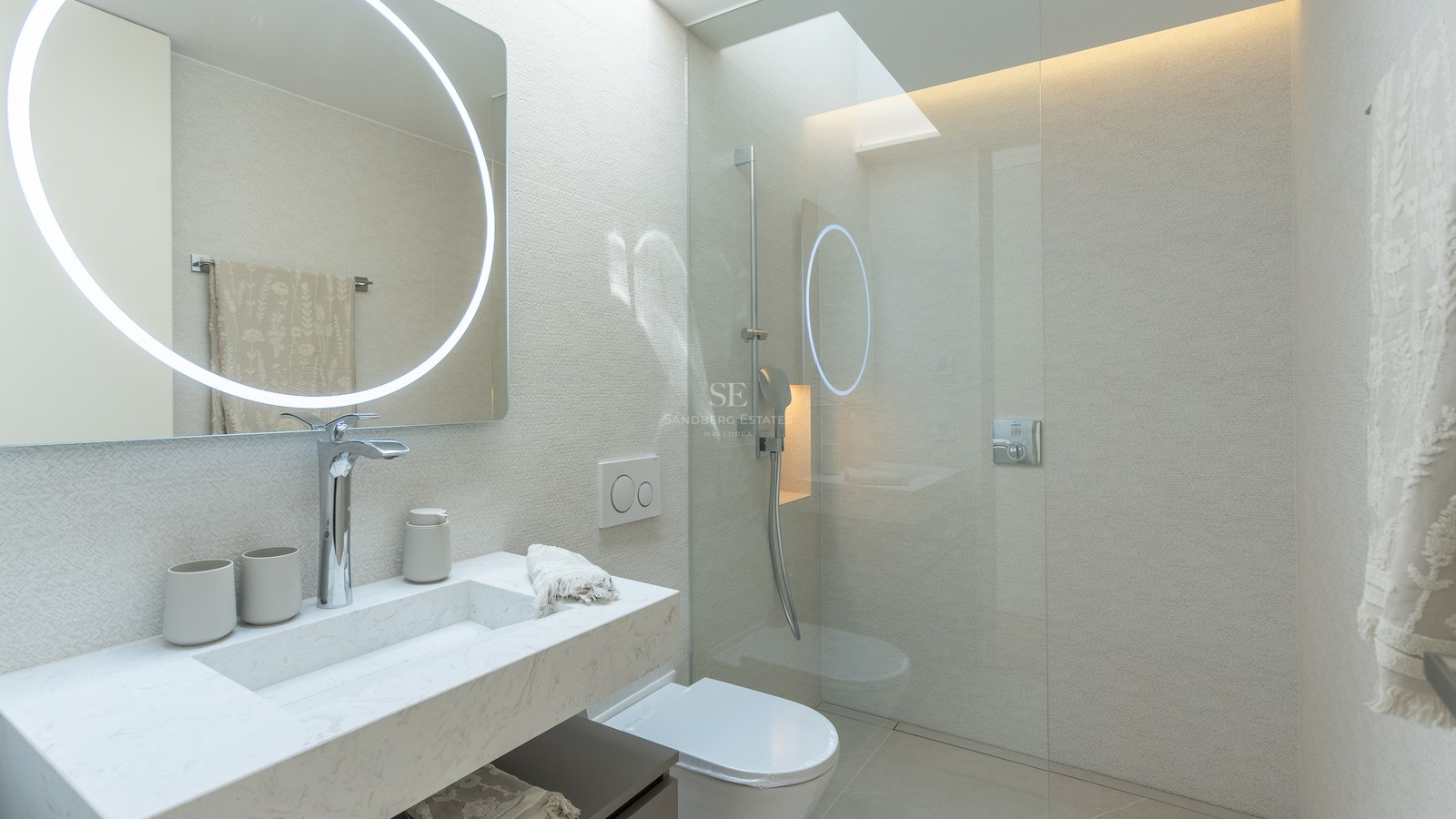 Modern bathroom featuring a circular LED mirror, marble vanity, and glass walk-in shower under a skylight.