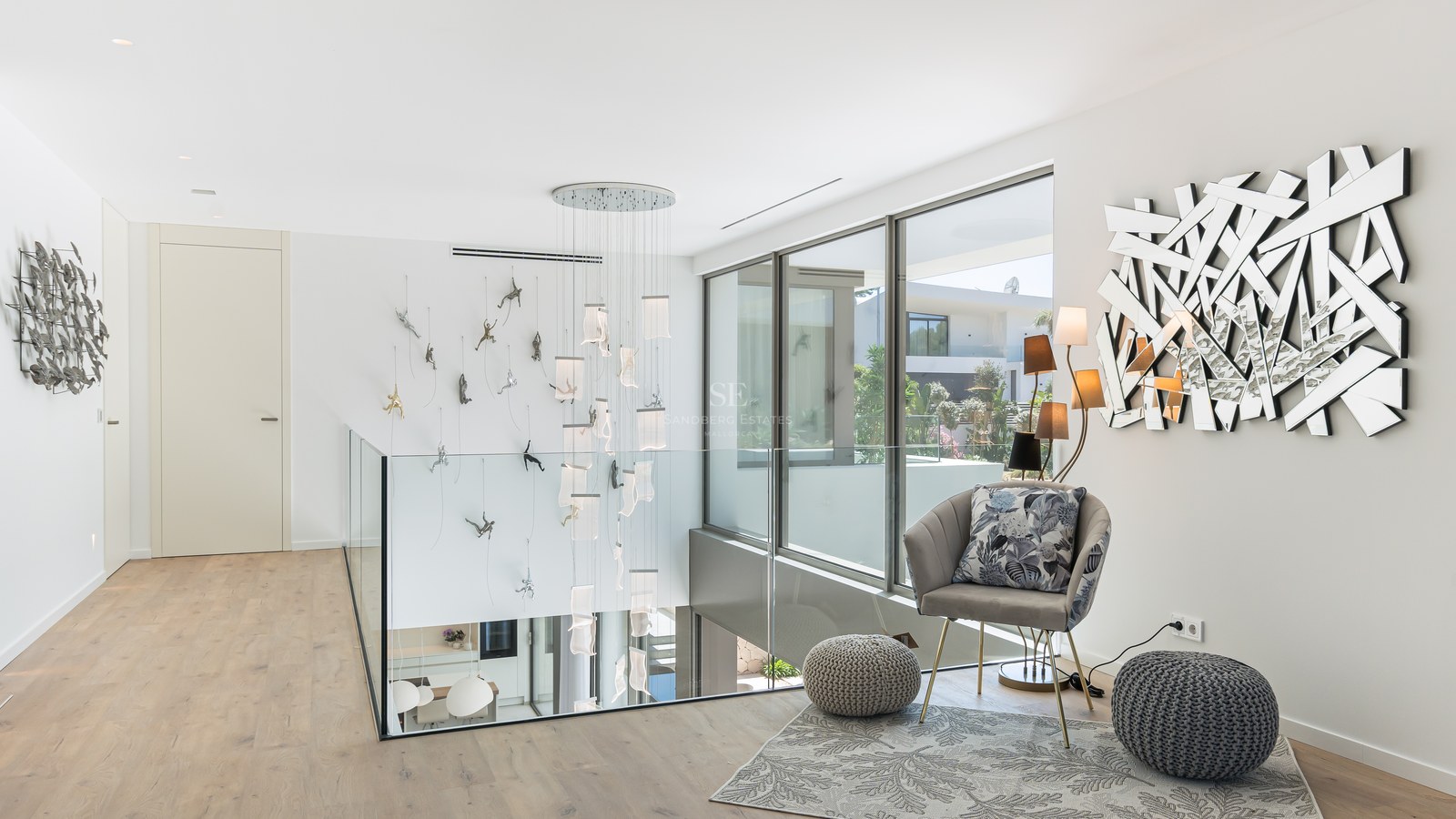A contemporary hallway landing with glass railings, a tall cascading chandelier, and floor-to-ceiling windows.