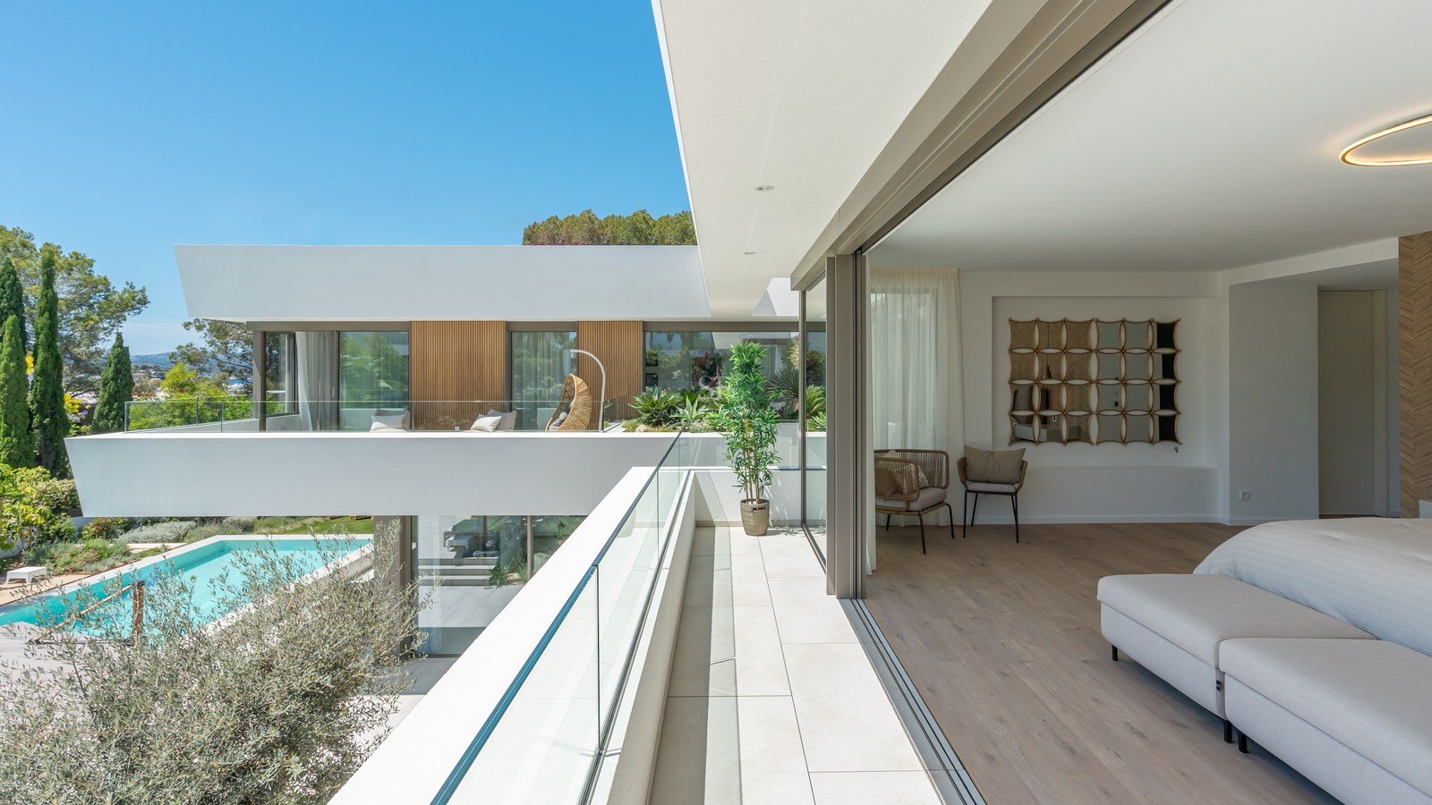 High-angle view of a modern master bedroom opening onto a white stone terrace with glass railings overlooking a pool.