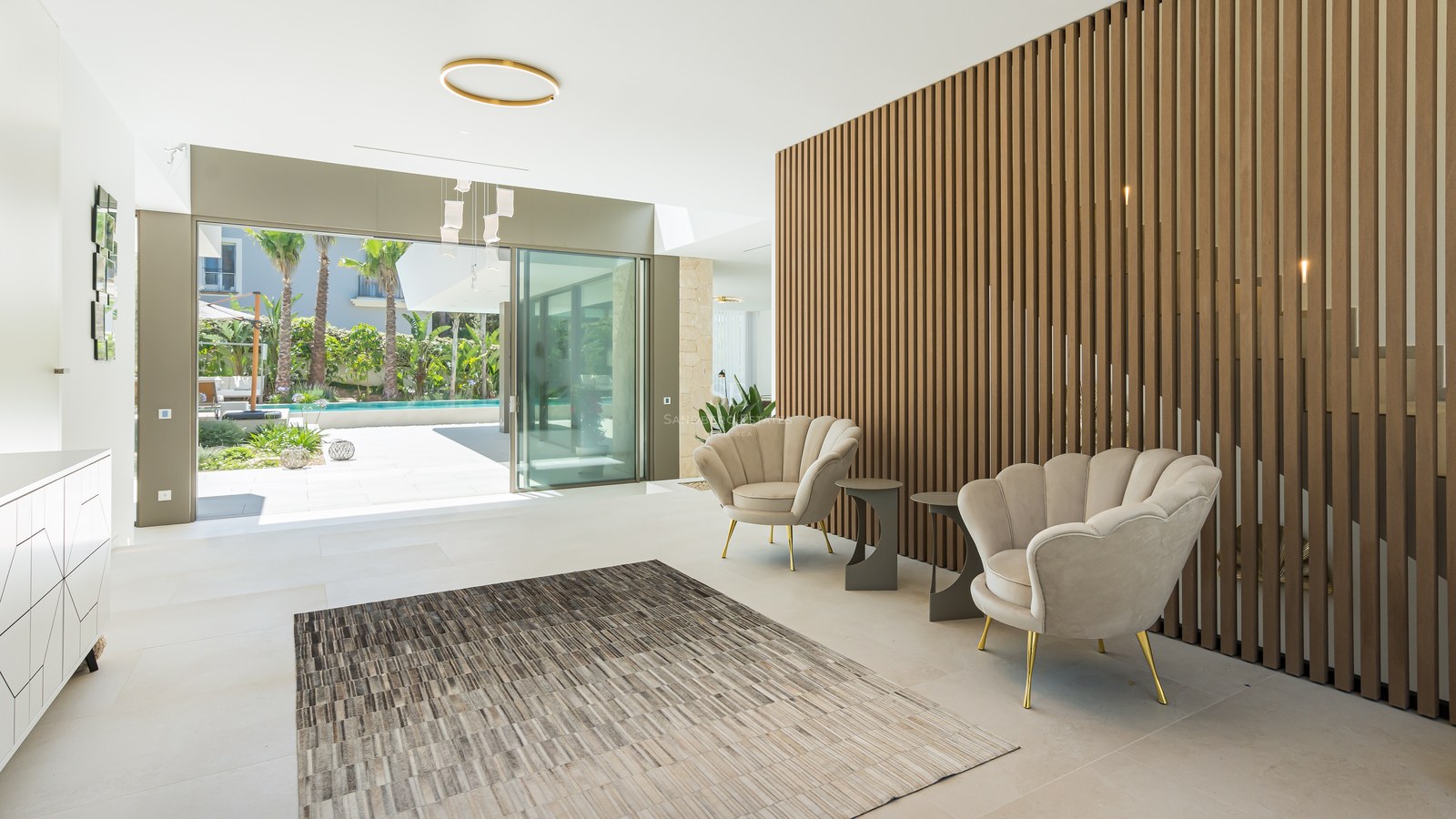 Modern lobby with wood slat wall, shell armchairs, and sliding glass doors opening to a pool and tropical garden.