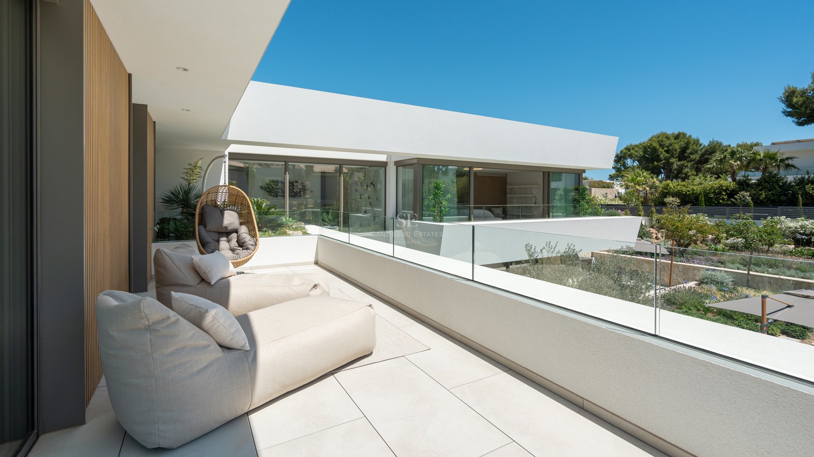 Spacious white terrace with beige lounge chairs, glass railings, and views of a landscaped garden under a clear blue sky.