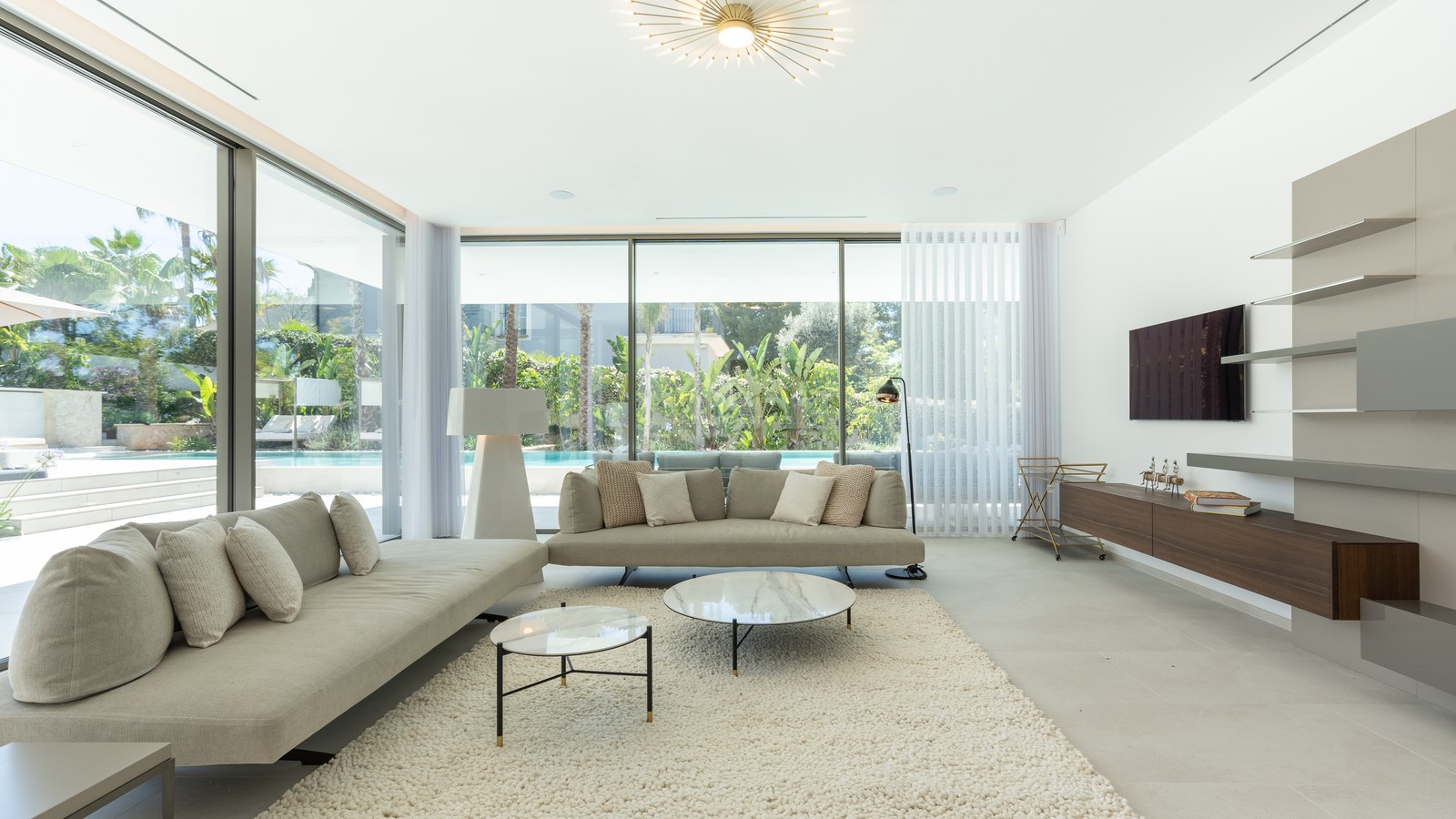 Contemporary living room featuring neutral sofas, marble coffee tables, and large windows overlooking a garden and pool.