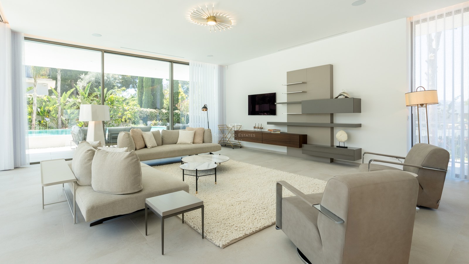 Contemporary living room with beige sofas, marble coffee tables, and floor-to-ceiling glass doors opening to a garden.