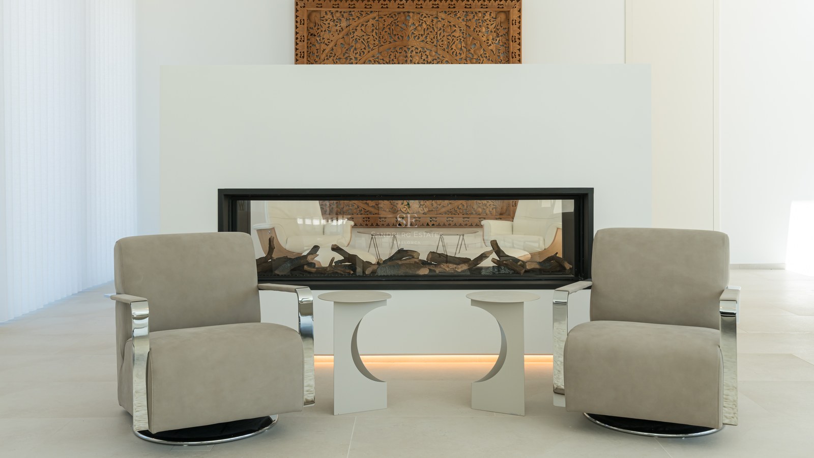 Two modern taupe armchairs facing a built-in linear fireplace with a hand-carved decorative wood panel above.