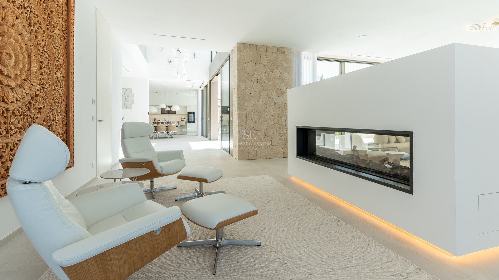 Two white lounge chairs facing a sleek glass fireplace in a bright, modern open-concept living space with stone pillars.