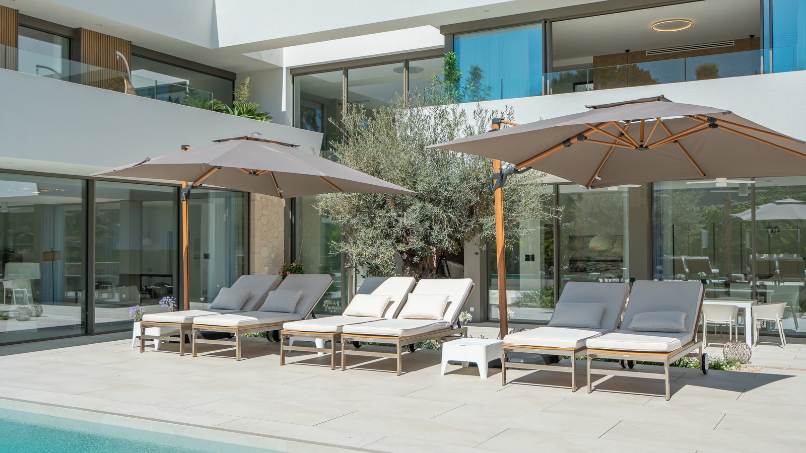 Luxury sun loungers and large umbrellas on a stone terrace beside a pool at a modern white villa.