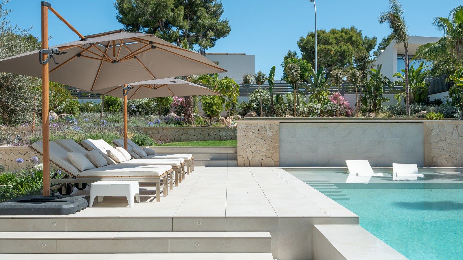 Modern swimming pool with in-water loungers and sun deck with parasols, surrounded by lush gardens.