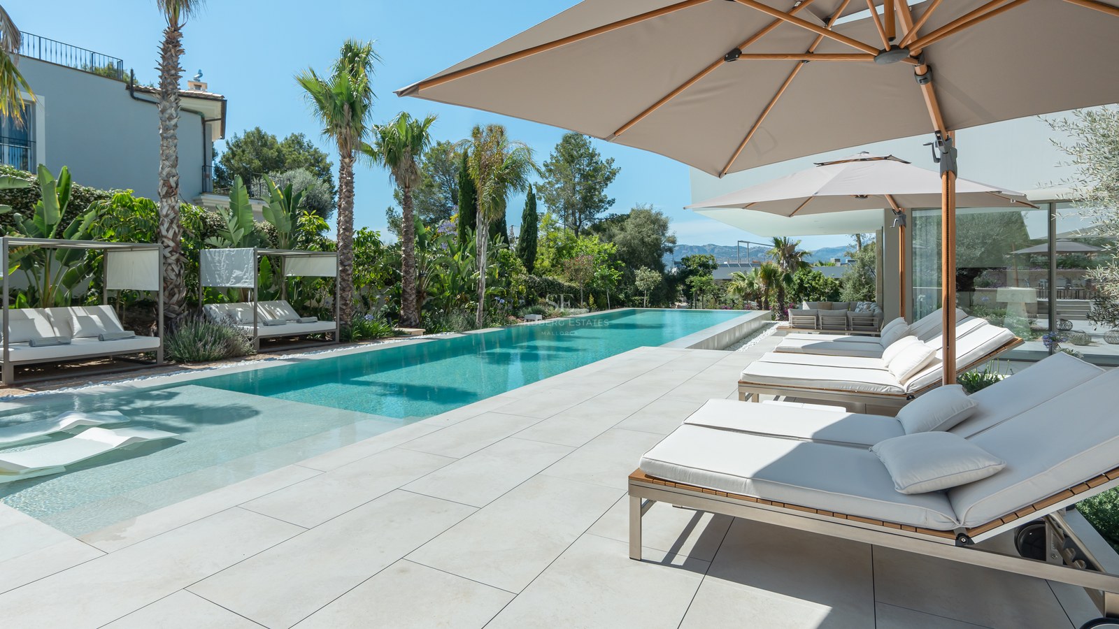 Luxury pool area with white sun loungers, beige umbrellas, and tropical palms under a clear blue sky.