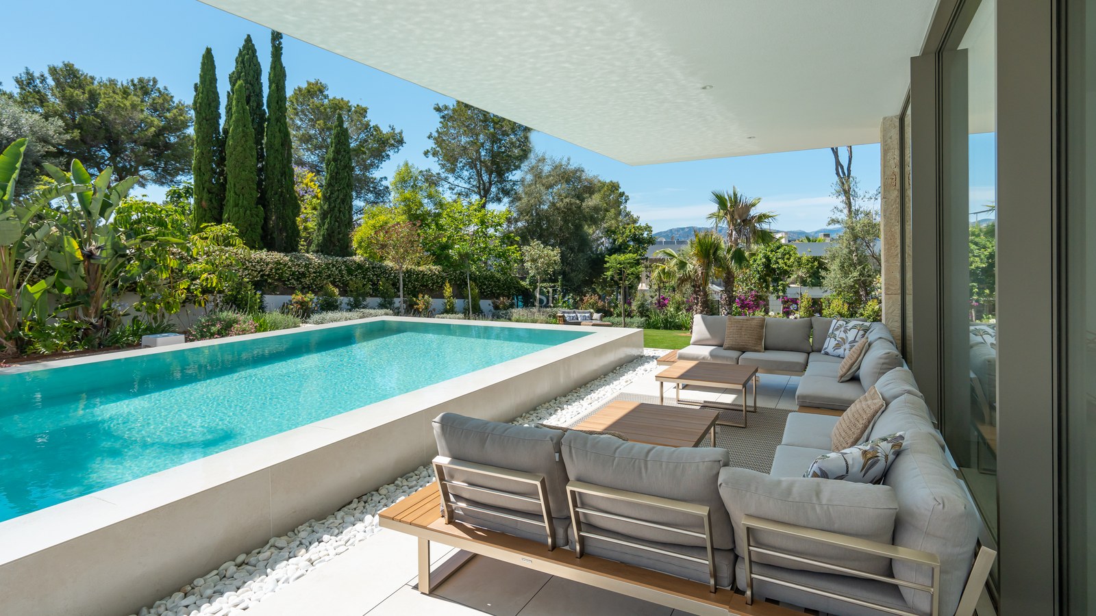 Raised infinity pool and gray lounge seating on a shaded terrace surrounded by lush Mediterranean greenery and mountain views.