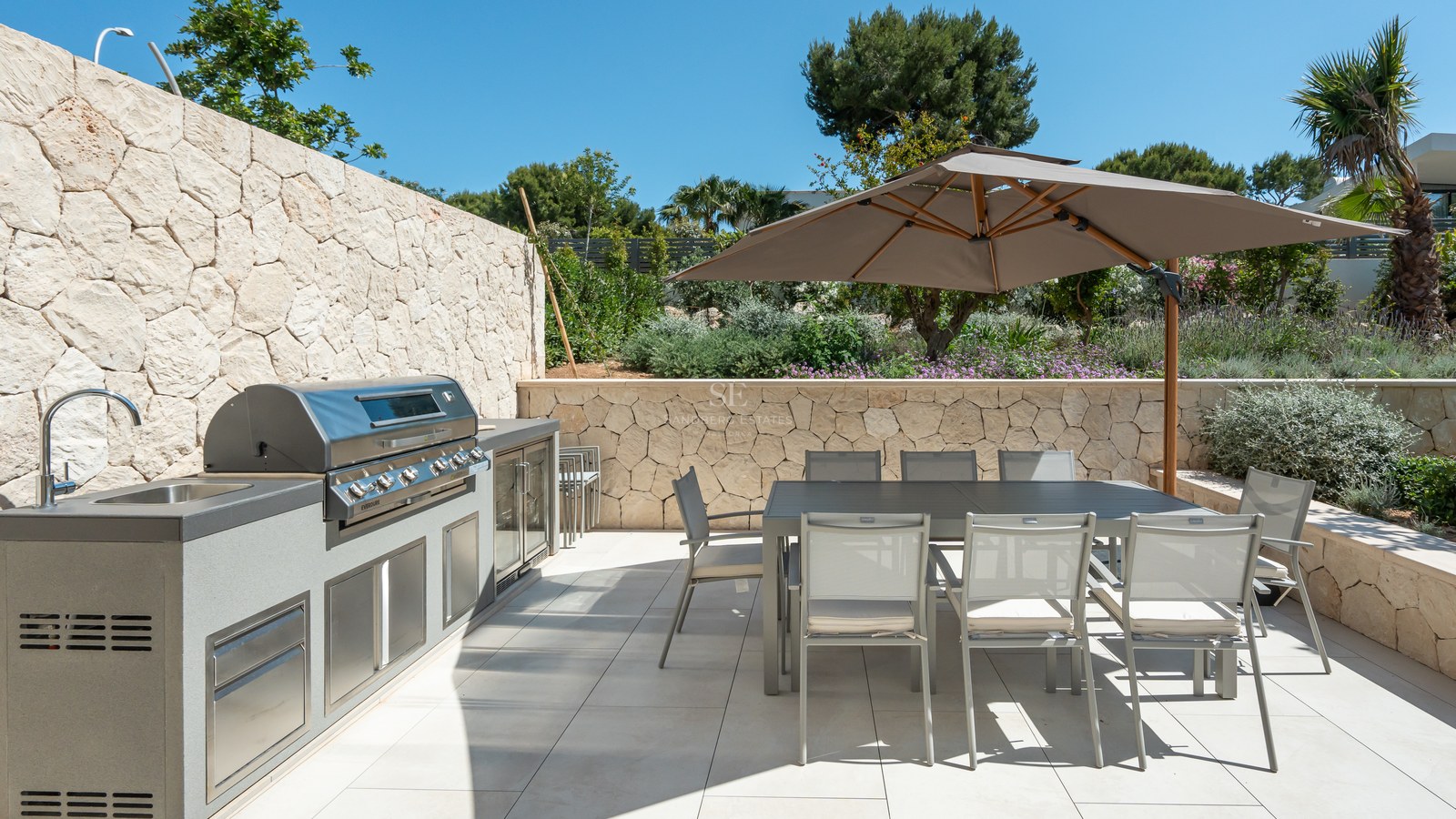 High-end outdoor kitchen with integrated BBQ and 8-seat dining table under a large umbrella on a stone-walled terrace.
