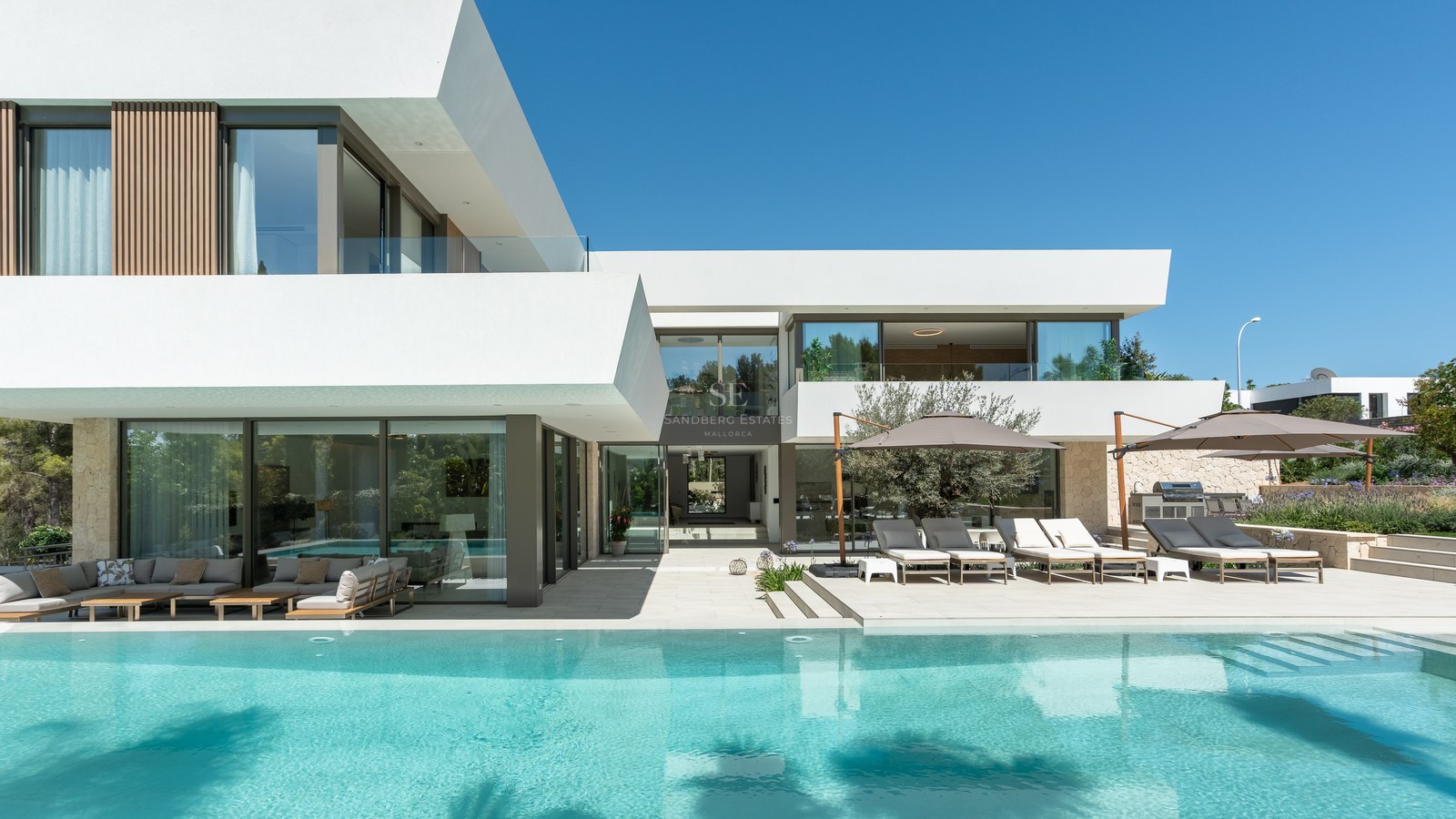 White modern villa with large swimming pool, sun loungers under umbrellas, and floor-to-ceiling glass windows.