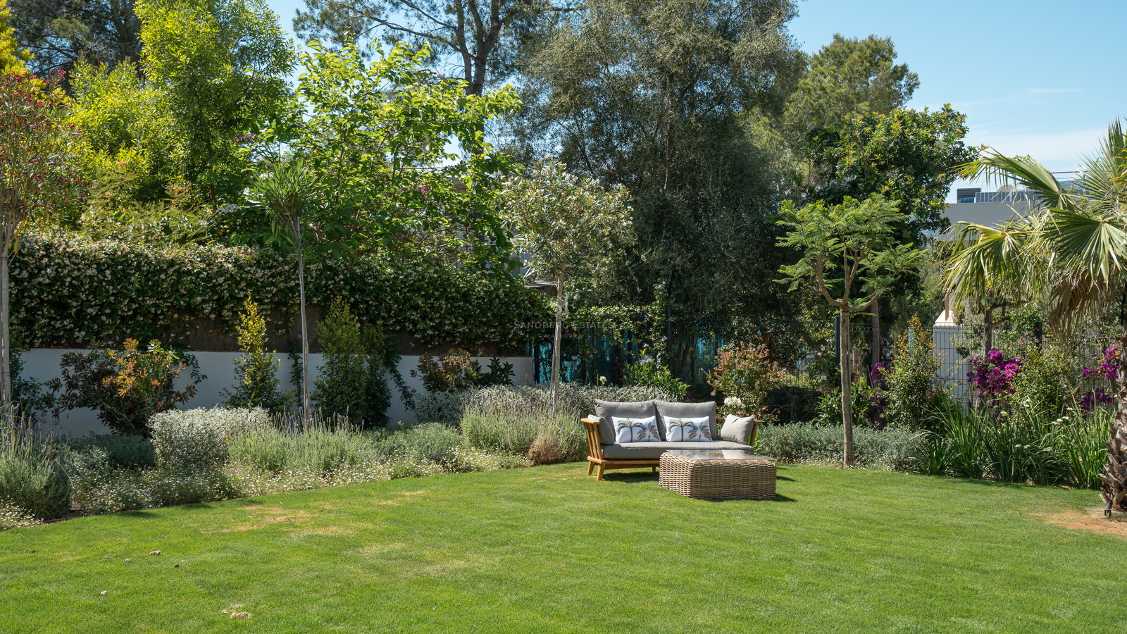 A lush green lawn surrounded by mature Mediterranean trees and flowering shrubs with a wooden sofa and wicker table.