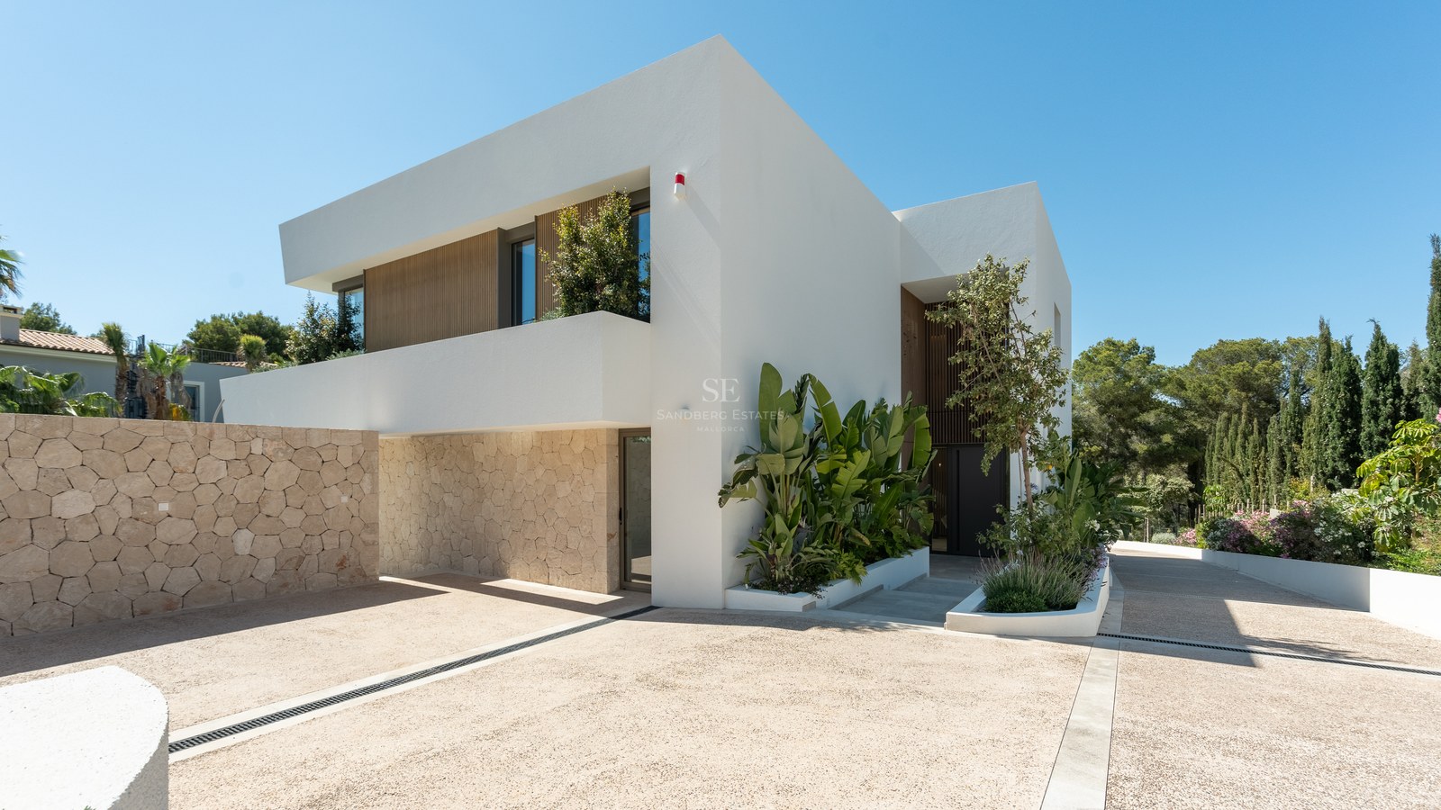 Contemporary two-story white villa with natural stone walls, tropical landscaping, and a spacious paved driveway under a blue sky.