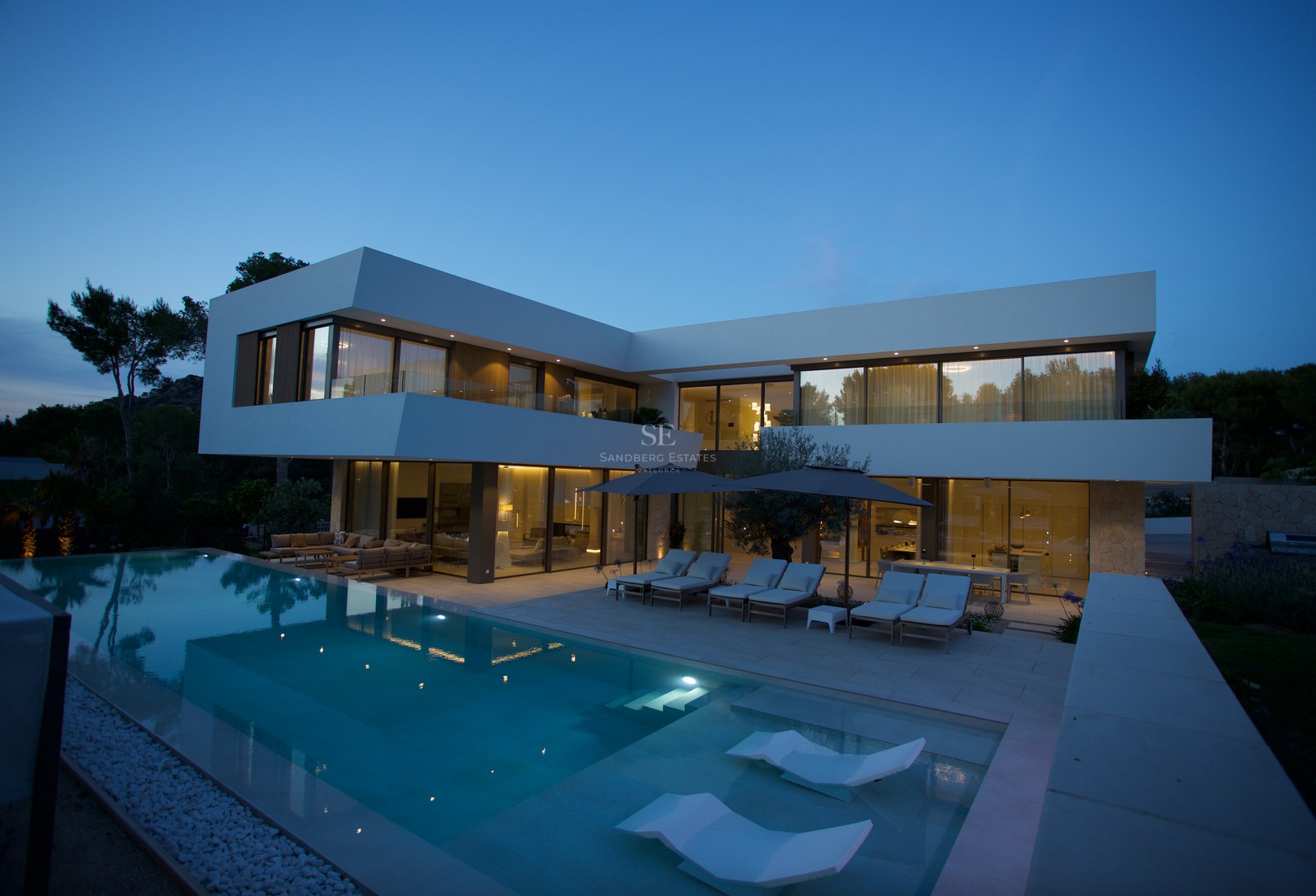 Contemporary two-story white villa with an illuminated infinity pool and submerged loungers under a twilight sky.