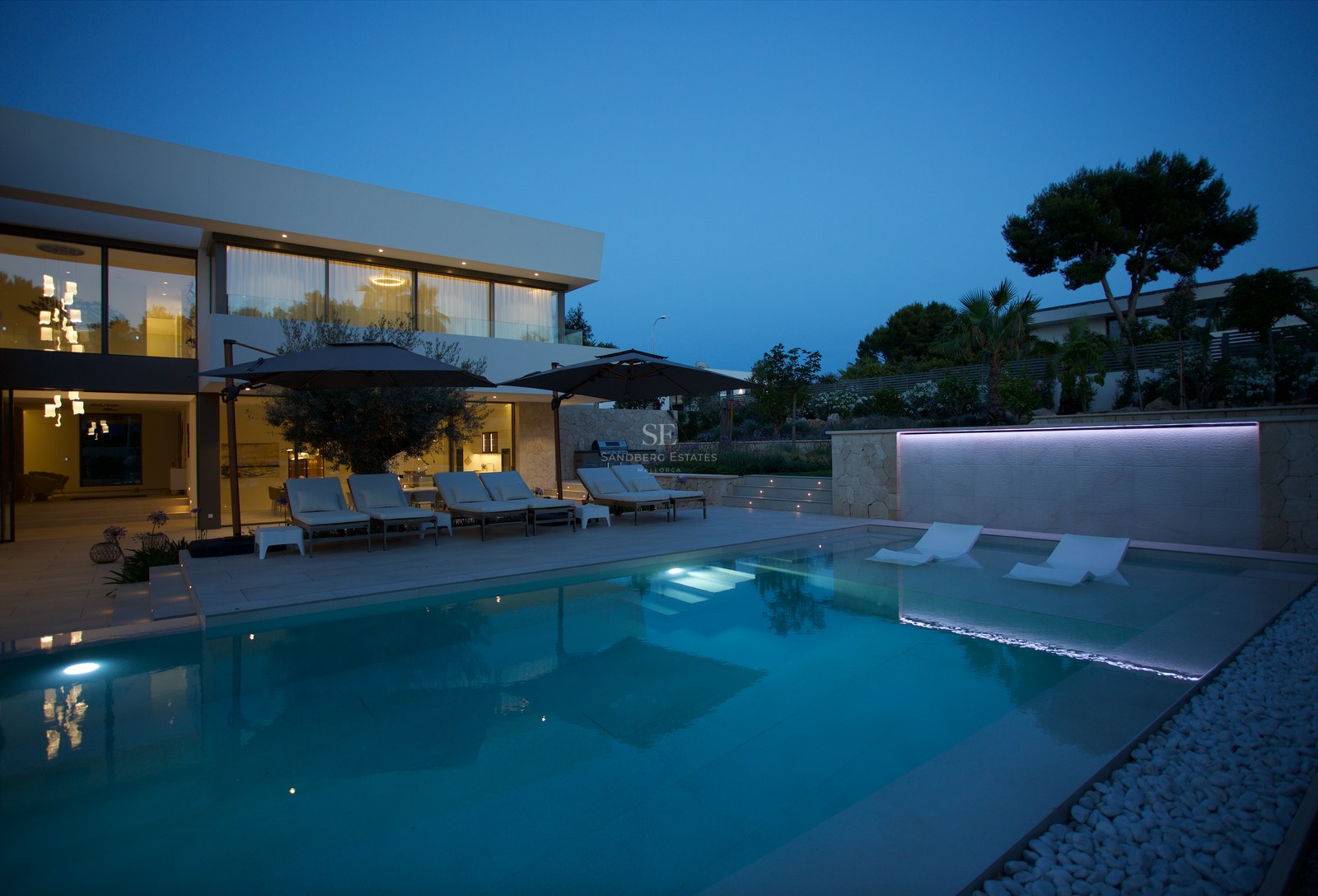 Modern villa exterior at dusk featuring a large swimming pool with integrated loungers and ambient lighting.
