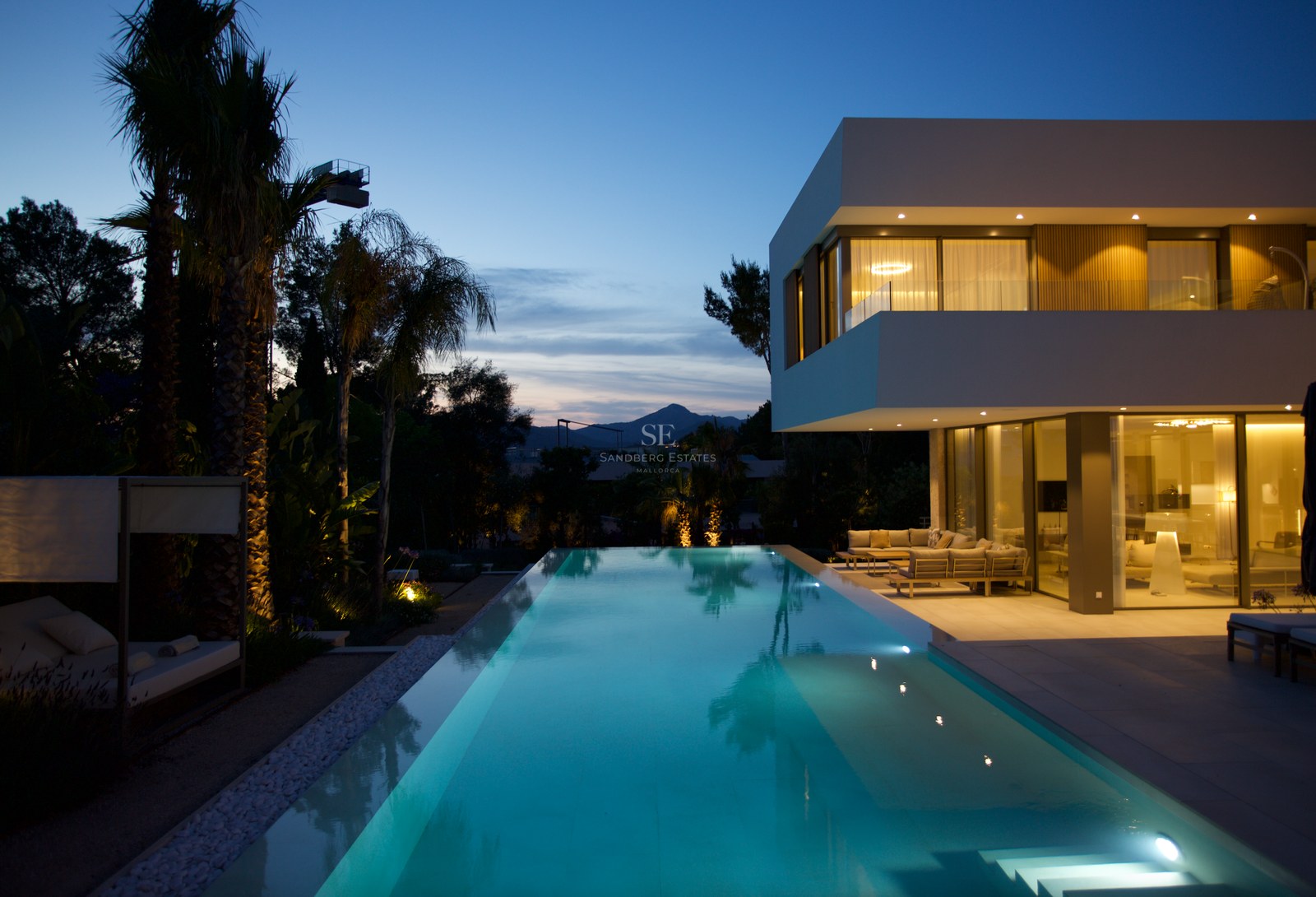 Contemporary two-story villa with a long swimming pool reflecting twilight sky and palm trees in a garden setting.