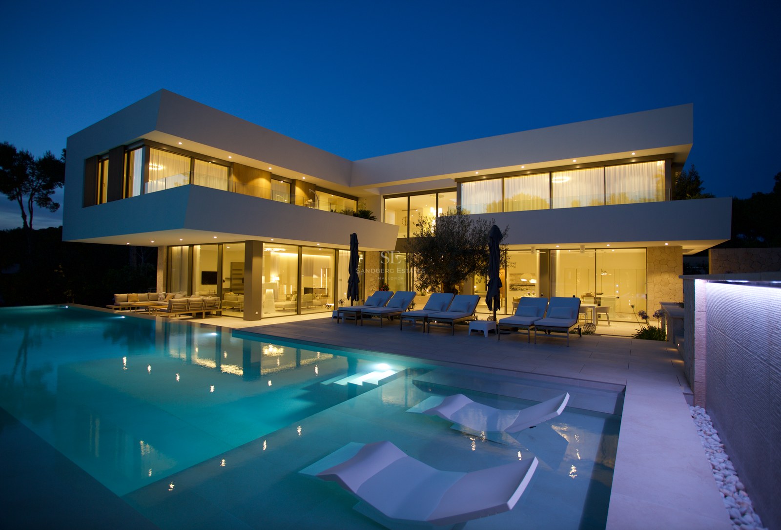Contemporary white two-story villa at night with a large lit swimming pool, submerged loungers, and minimalist facade.
