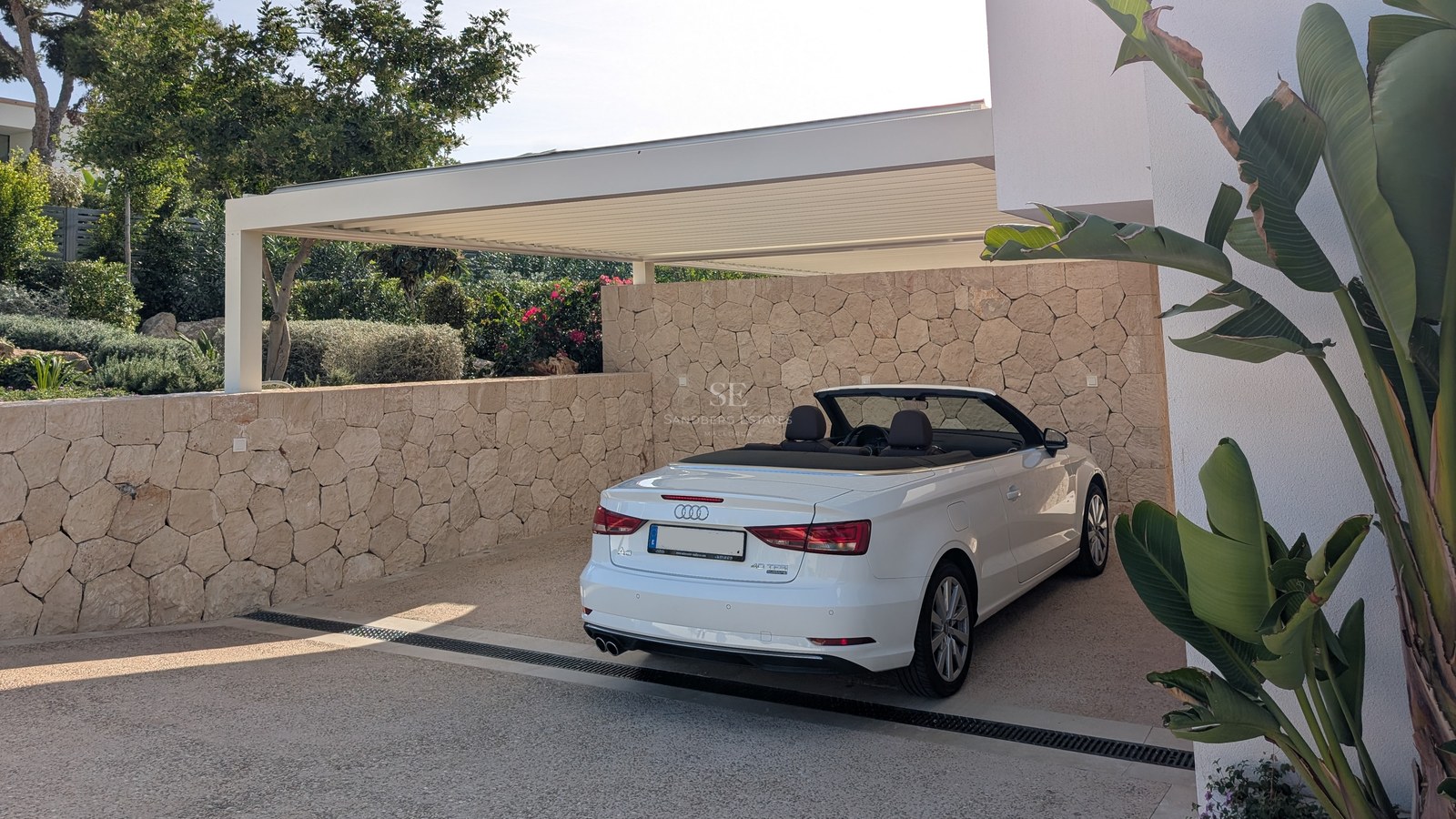 A white convertible parked under a modern louvered pergola, surrounded by natural stone walls and lush greenery.