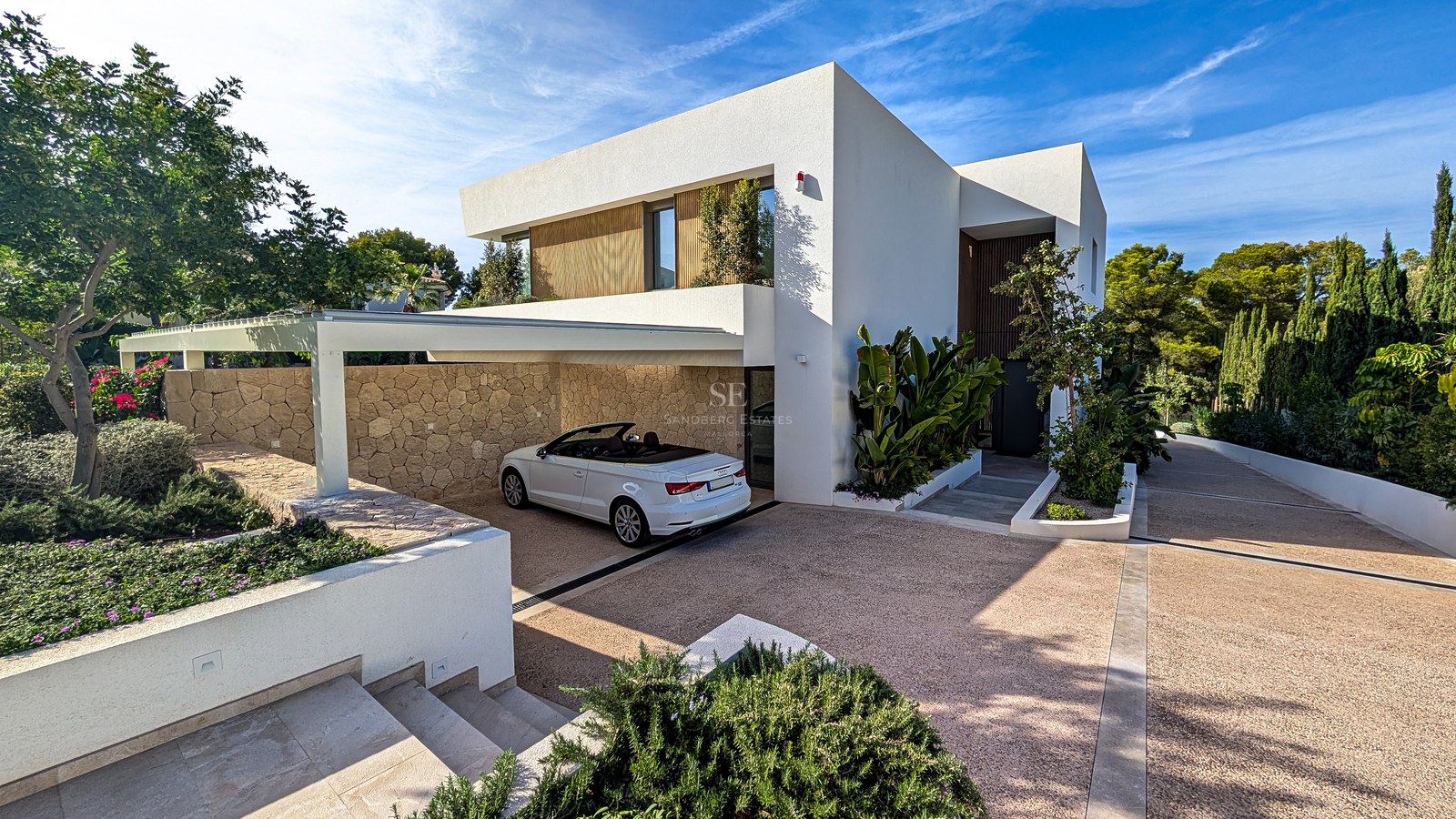 Modern white villa with a white convertible parked in a natural stone-walled carport under a clear blue sky.