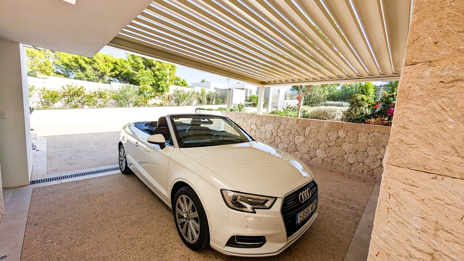White convertible car parked under a modern slatted pergola next to a natural stone wall and garden greenery.
