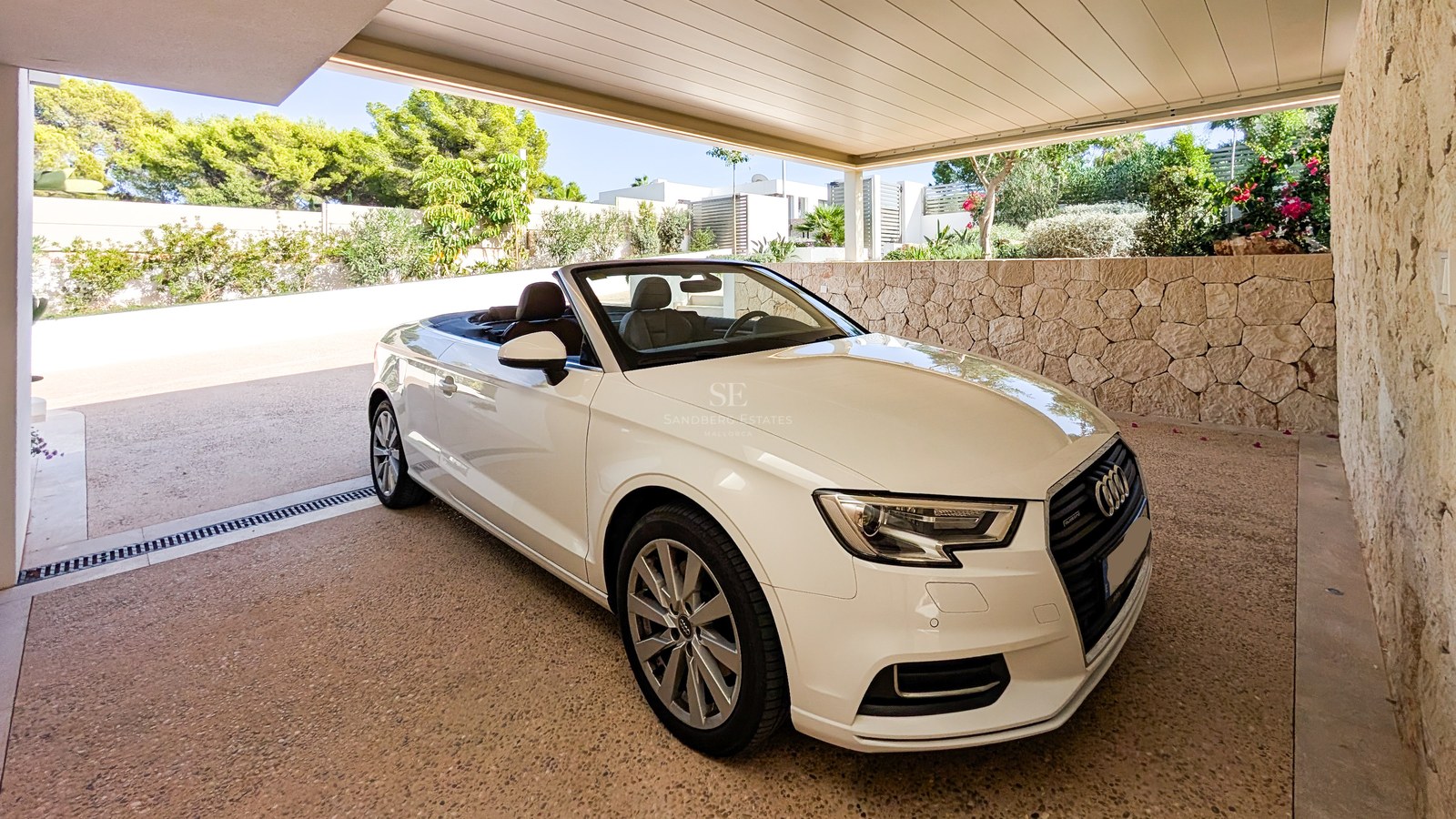 White convertible parked in a modern covered carport with natural stone walls and lush Mediterranean landscaping.