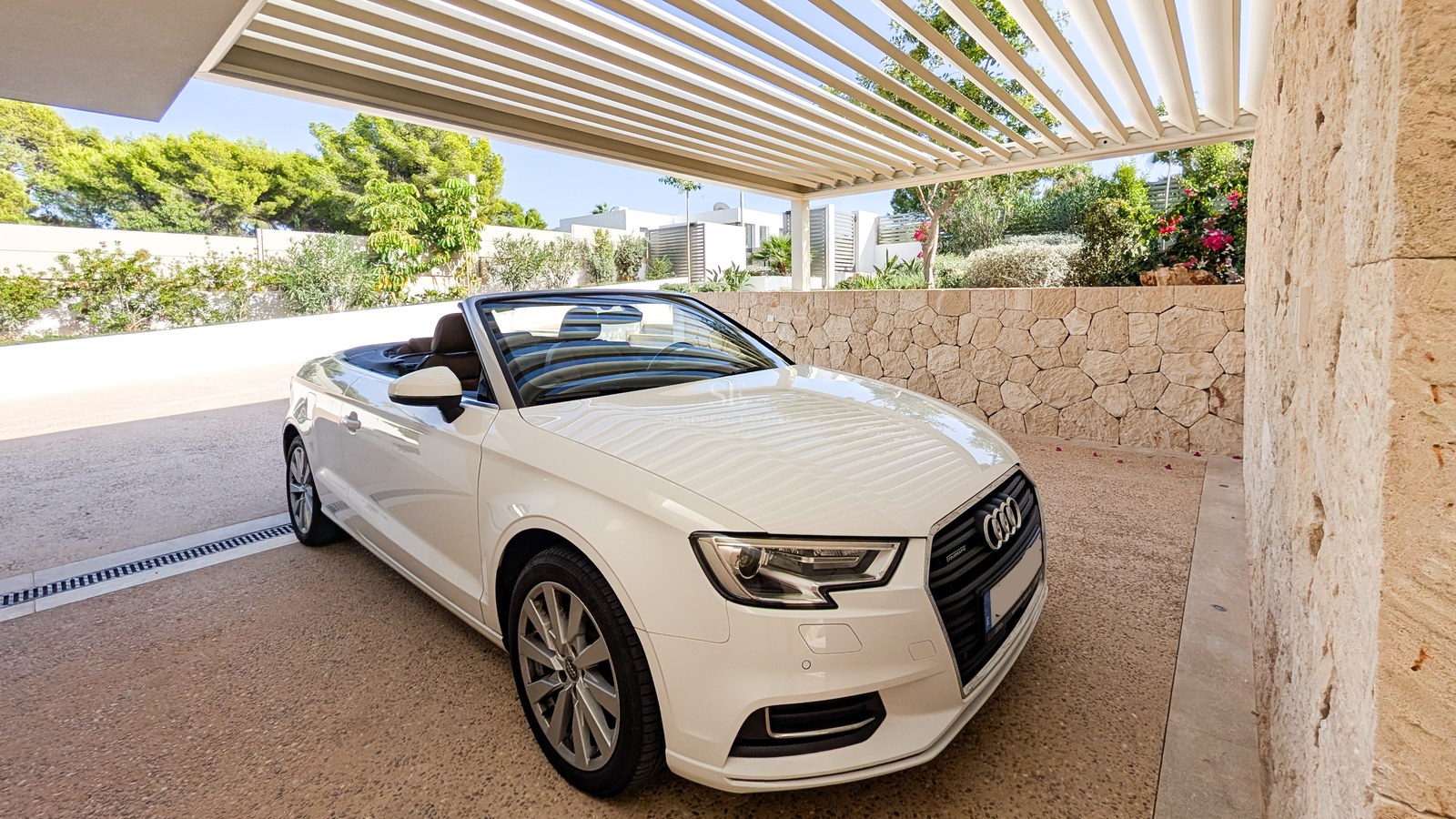 White convertible car parked under a modern slatted pergola next to a natural stone wall and lush garden.