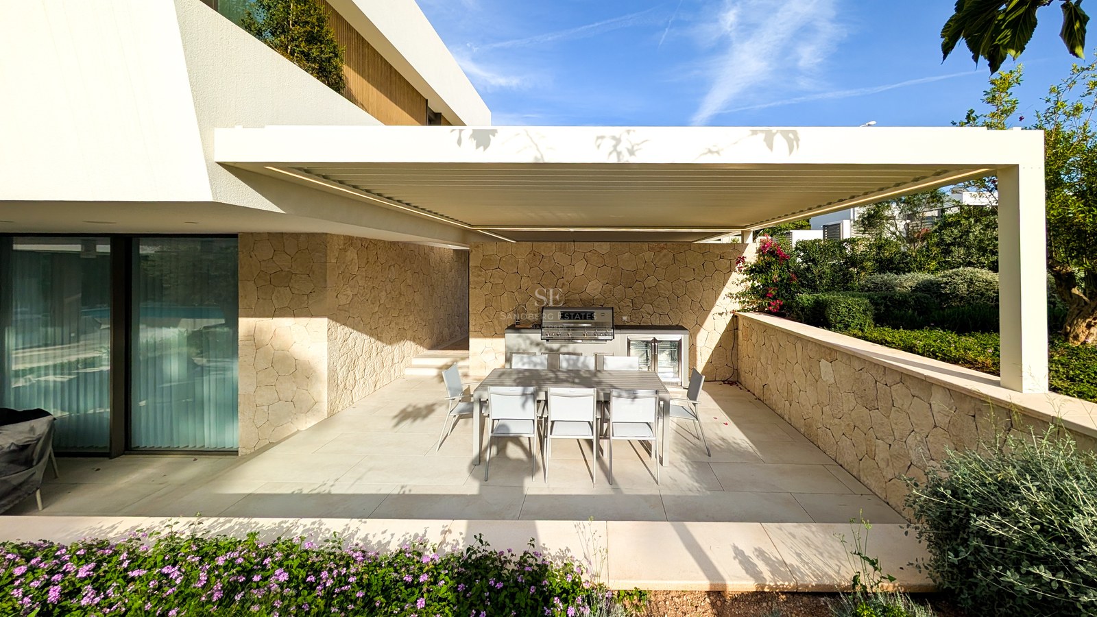 Covered outdoor kitchen with stainless steel grill and dining table for eight against natural stone walls.
