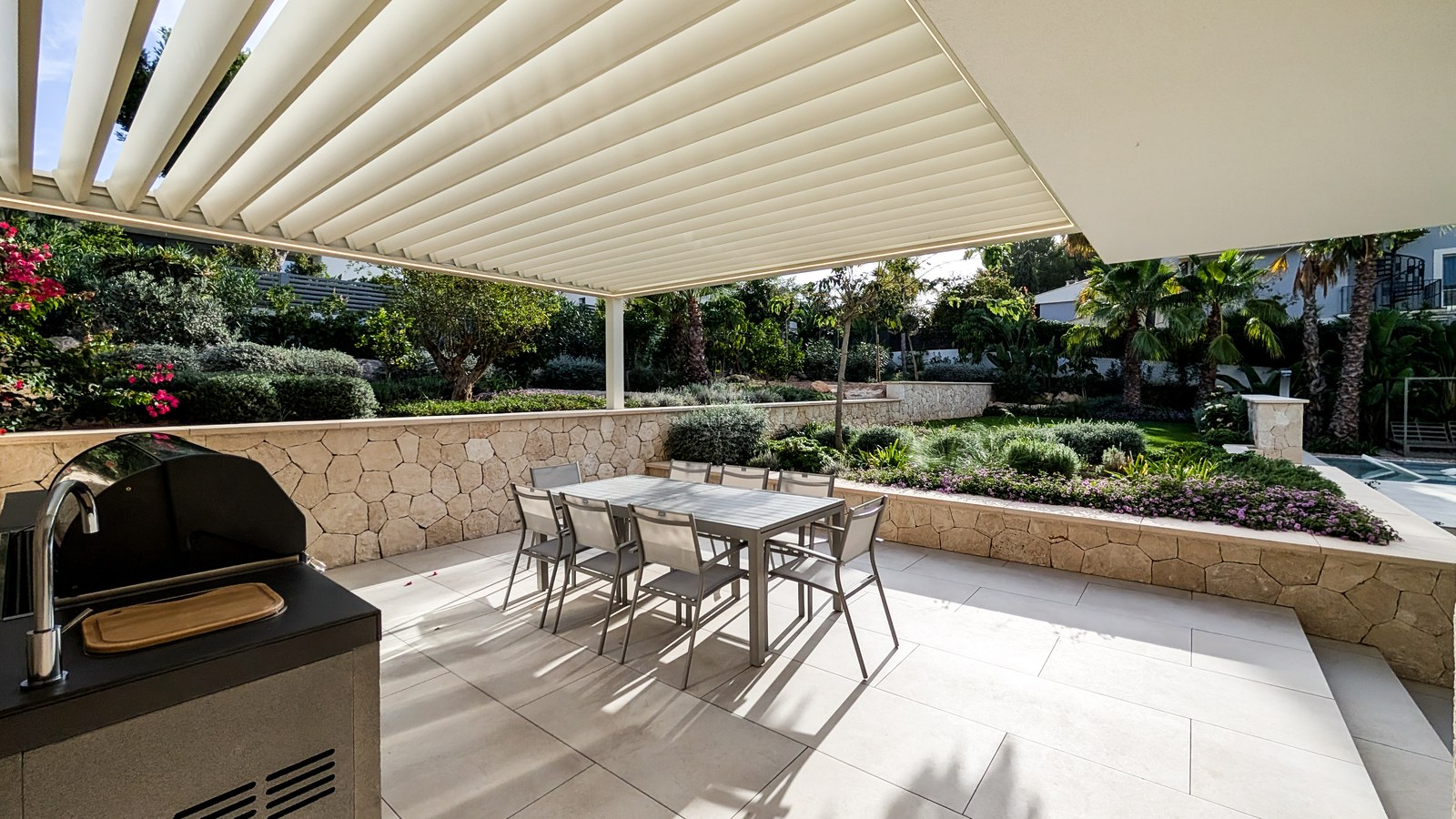 Modern terrace with a large dining table under a white pergola, featuring an outdoor kitchen and stone walls.